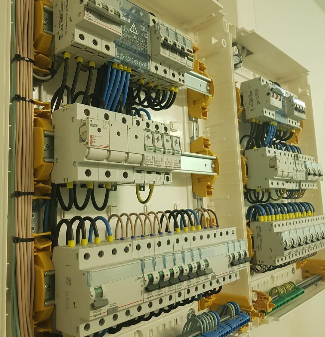 Rozdzielnica elektryczna z zainstalowanymi bezpiecznikami i okablowaniem w kolorach niebieskim, brązowym i czarnym. Widoczne elementy marki Legrand w jasnej obudowie.