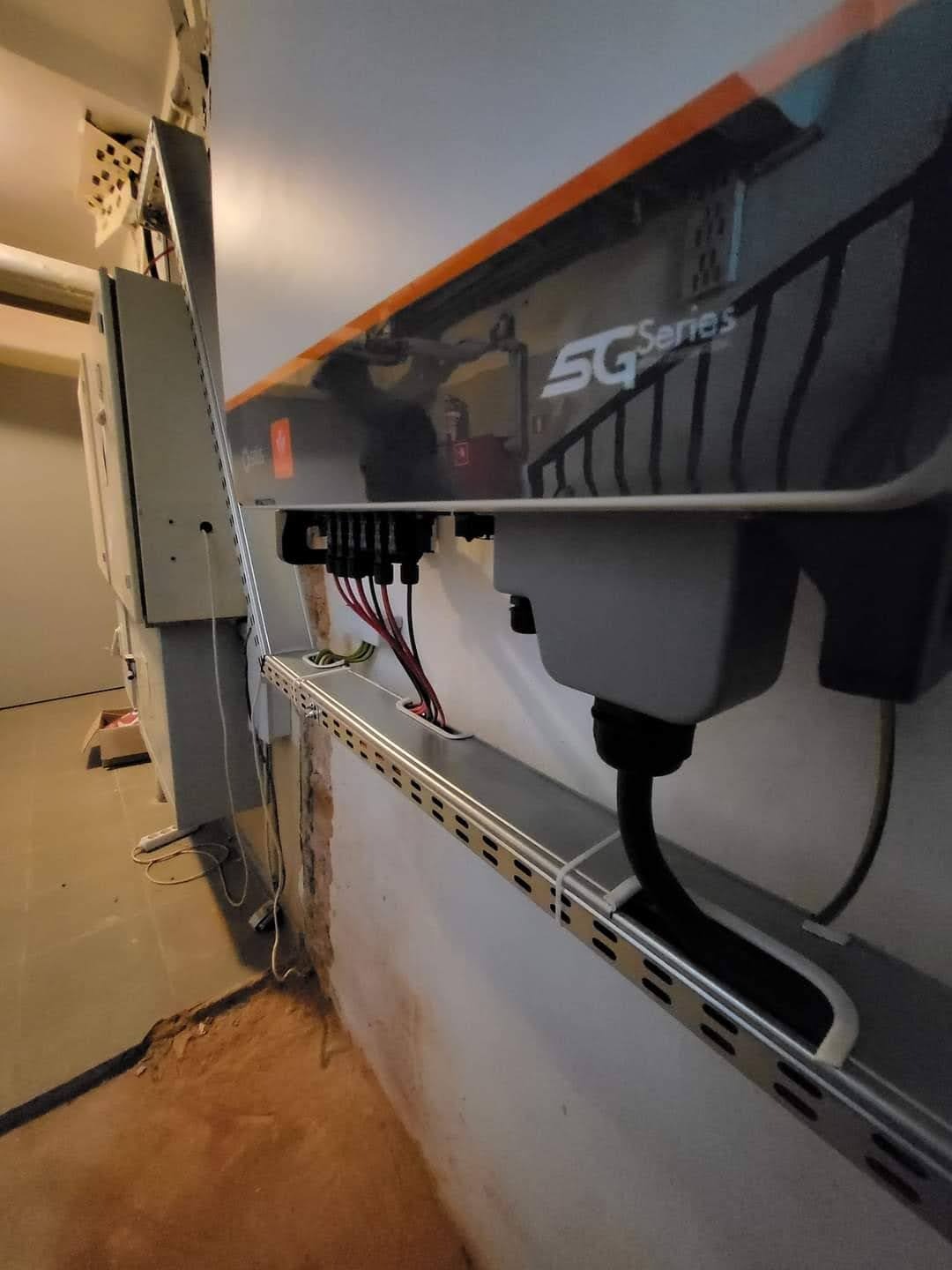 Szary inwerter 5G Series zamontowany na ścianie z widocznymi przewodami i metalową listwą. Instalacja w surowym pomieszczeniu, widoczny fragment podłogi i ściany.