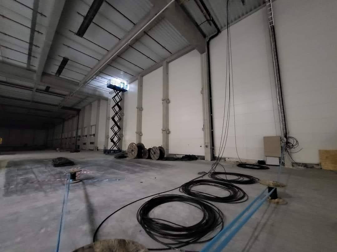 Wnętrze hali w trakcie instalacji elektrycznej. Widoczna podnośnik nożycowy, kable w kręgach, skrzynka elektryczna na ścianie i elementy instalacji pod sufitem.