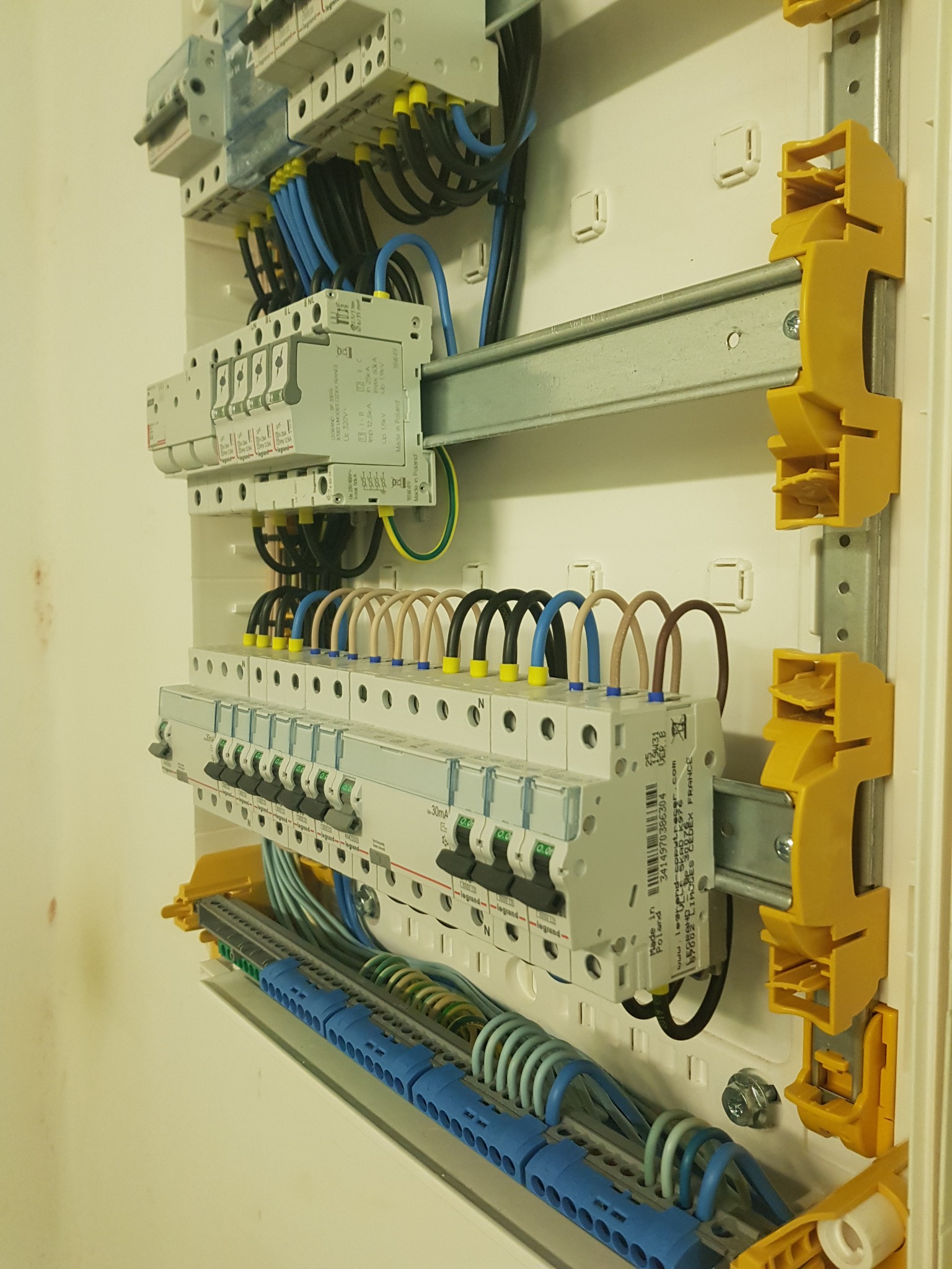 Rozdzielnica elektryczna z widocznymi bezpiecznikami, przewodami w różnych kolorach i elementami montażowymi w kolorze żółtym i niebieskim.