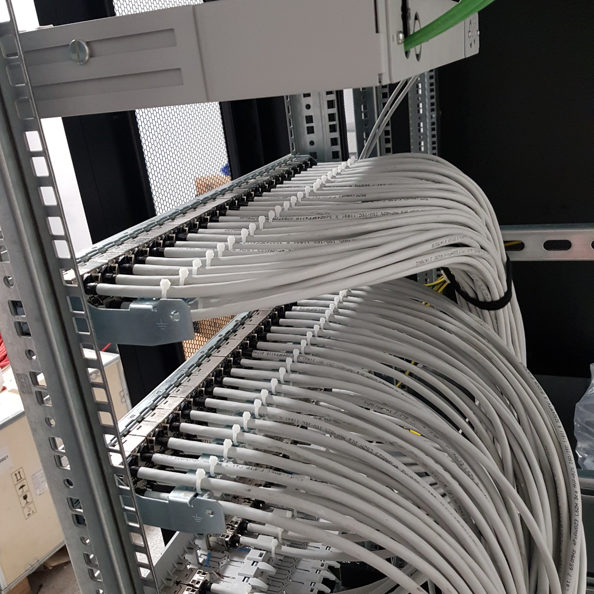 Uporządkowane kable sieciowe w szafie rack, precyzyjnie ułożone i zabezpieczone. Widoczne oznaczenia na kablach i metalowa konstrukcja szafy.