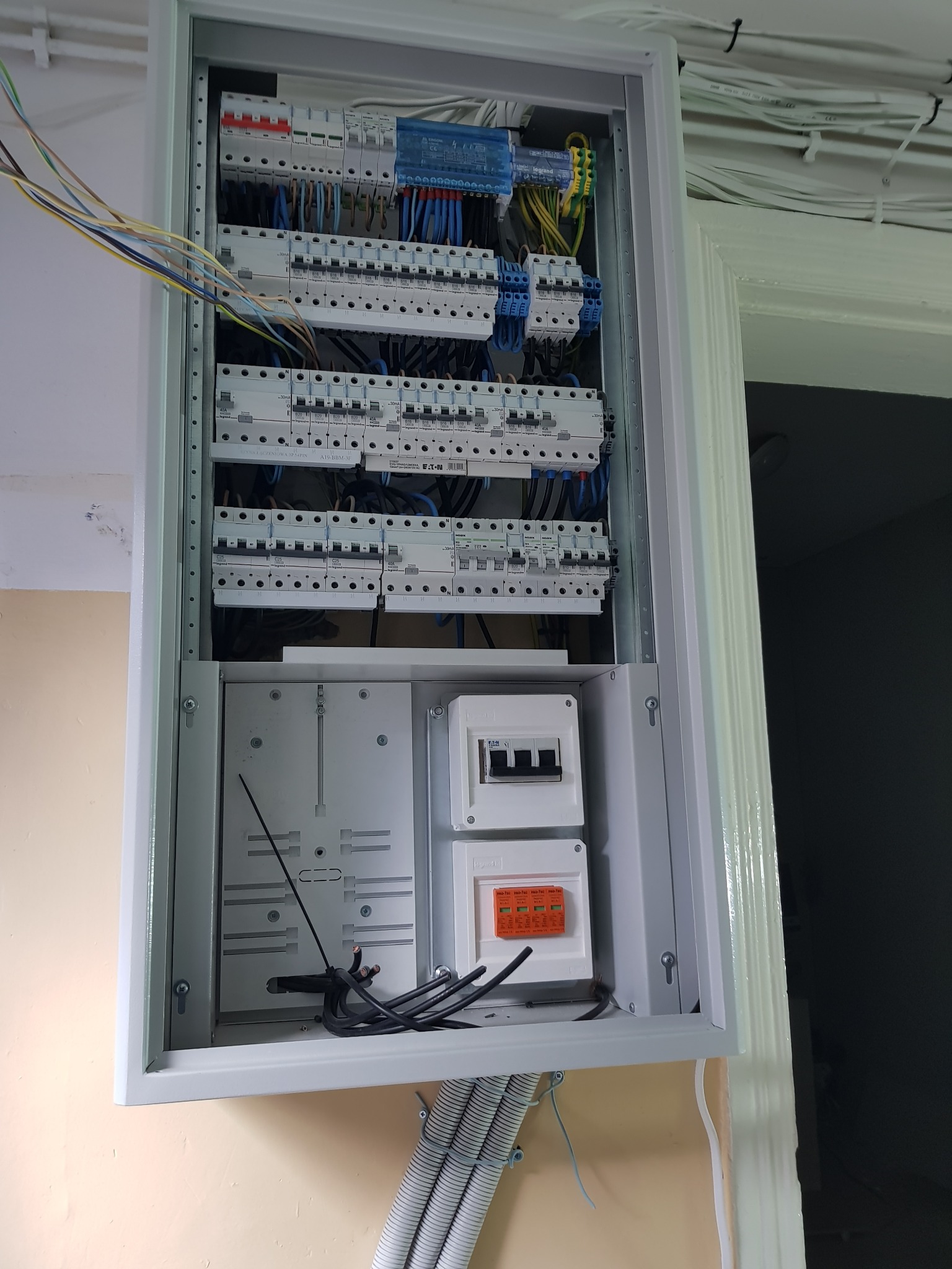 Otwarta, szara skrzynka elektryczna z zainstalowanymi zabezpieczeniami i przewodami. Widoczne kable wchodzące do skrzynki. Całość zamontowana na ścianie.