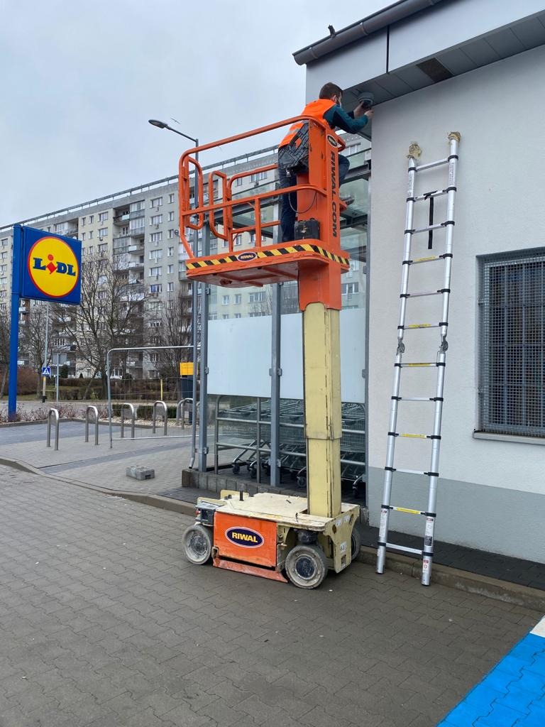 Pracownik na podnośniku koszowym dokonuje naprawy/montażu na elewacji budynku z logo Lidl. Obok drabina. Ujęcie z dołu, na tle bloku mieszkalnego.