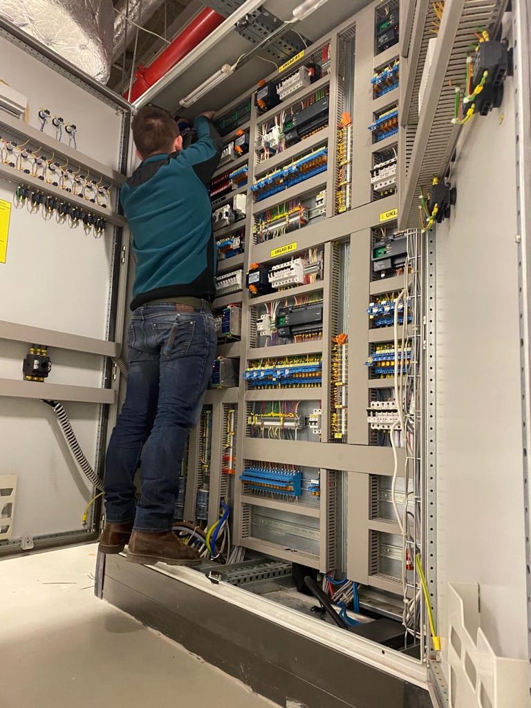 Elektryk na podeście, w trakcie pracy przy rozbudowanej szafie sterowniczej z licznymi przewodami i elementami elektrycznymi. Widok z dołu, wnętrze pomieszczenia.