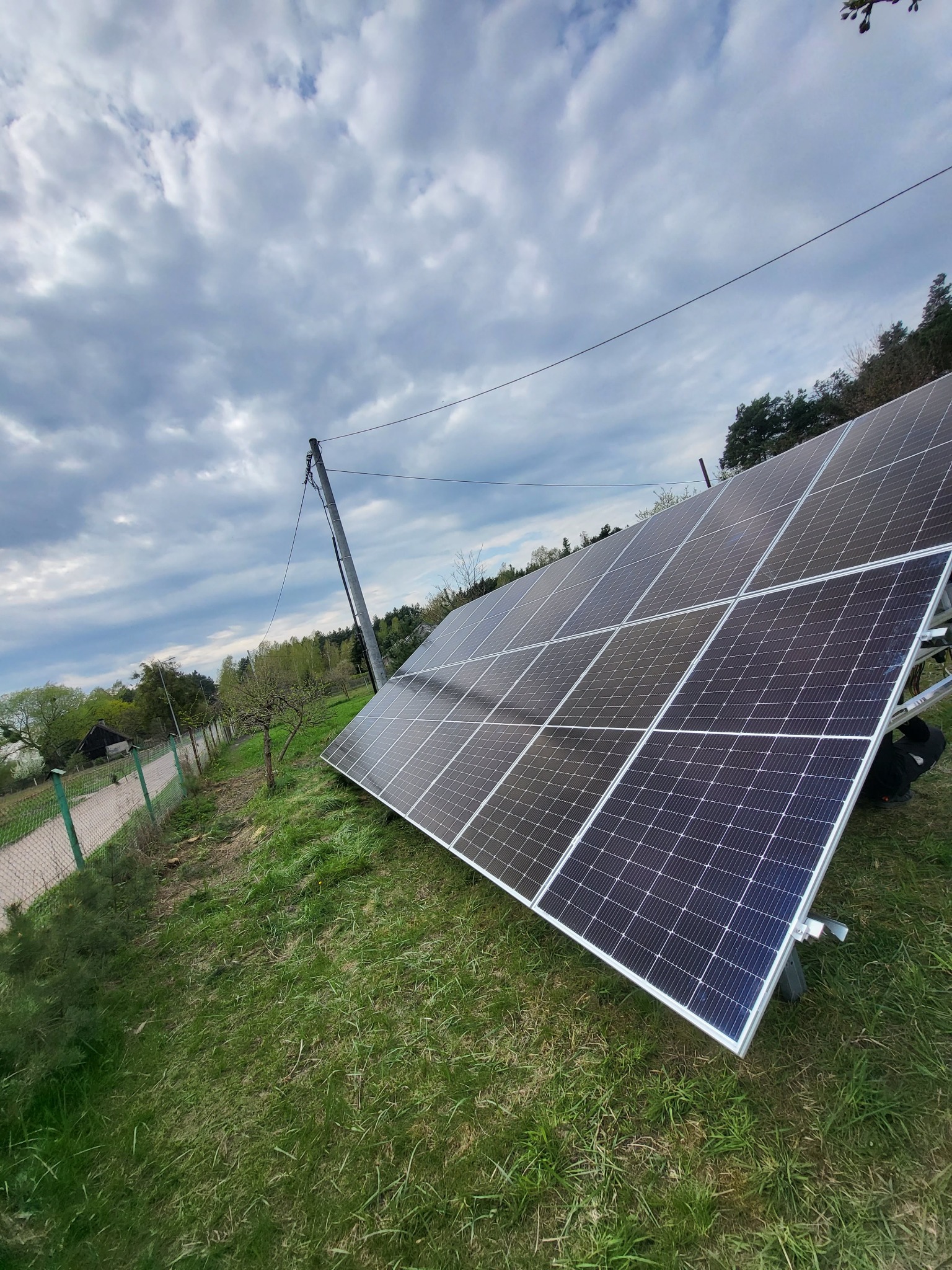Panel fotowoltaiczny na trawie, widok z dołu, pochmurne niebo. Instalacja przy ogrodzeniu, słup elektryczny w tle. Układ paneli w srebrnej ramie.