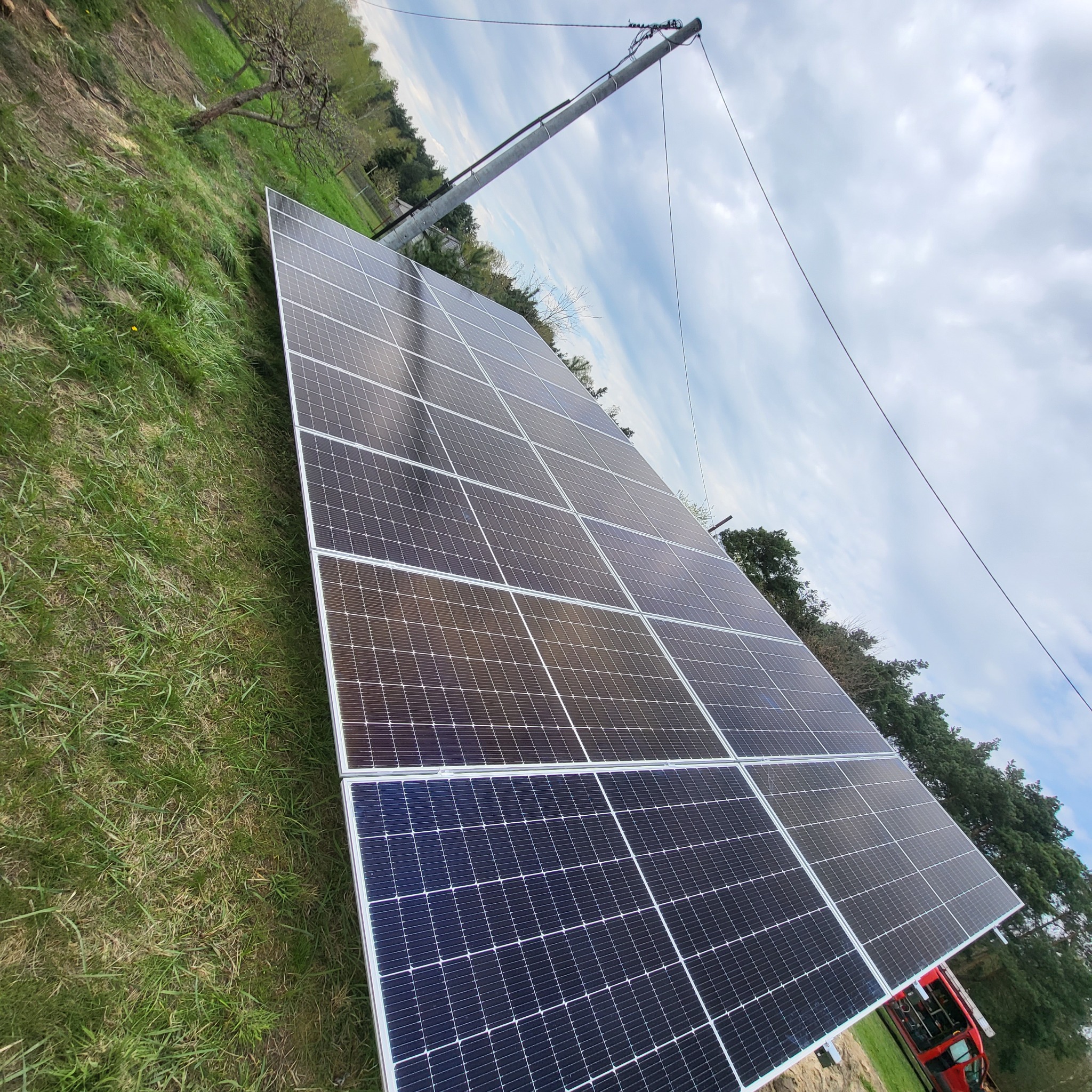 Panel fotowoltaiczny na trawie, widok z dołu. Słup energetyczny w tle. Instalacja paneli słonecznych w otoczeniu zieleni i błękitnego nieba z chmurami.