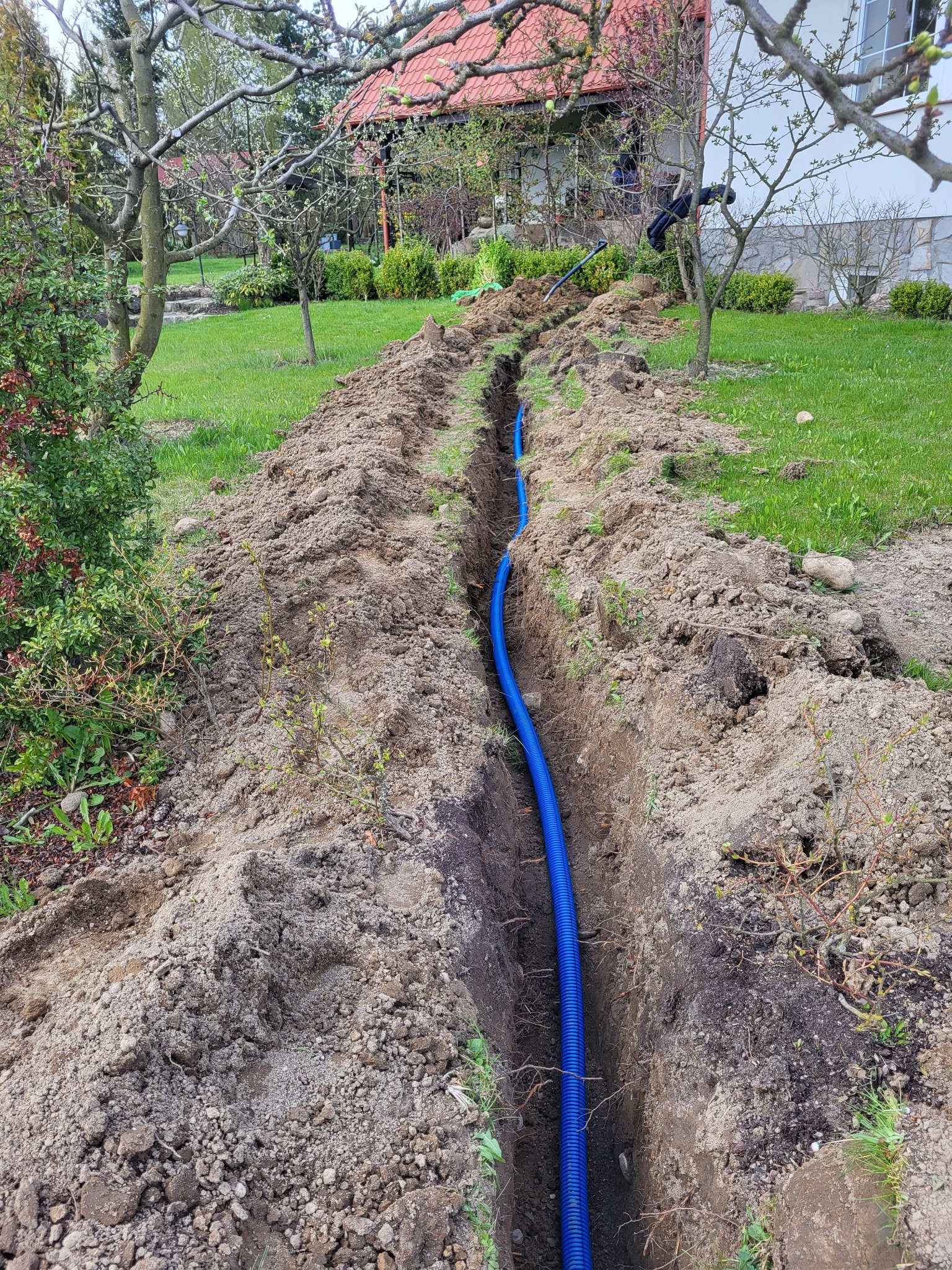 Niebieska rura karbowana w wykopie na trawniku, prawdopodobnie instalacja podziemna. Widok z góry na tle domu i drzew.