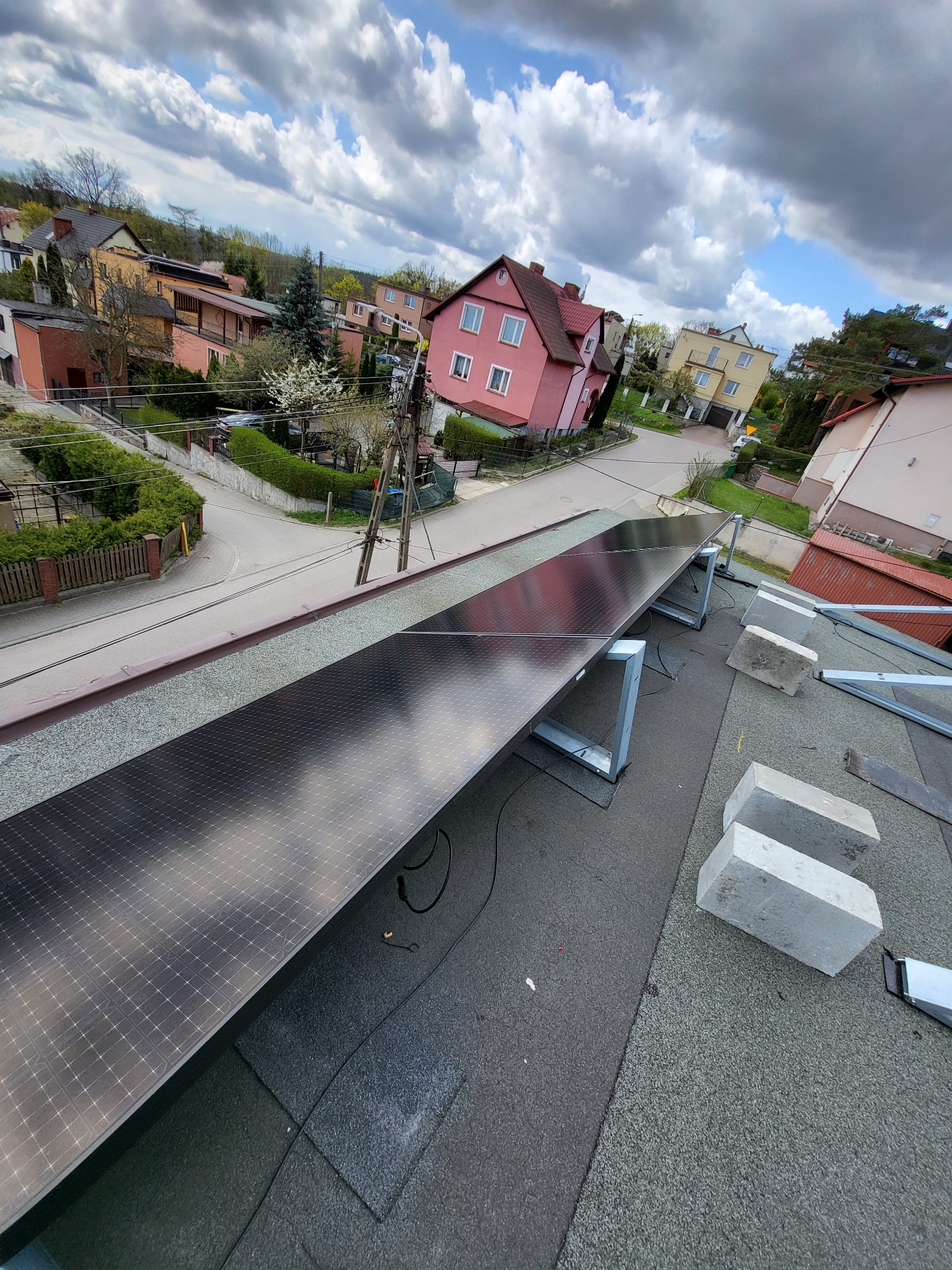 Montaż paneli fotowoltaicznych na dachu domu jednorodzinnego, widok z góry na częściowo zamontowaną instalację, w tle zabudowa mieszkalna i fragment nieba.