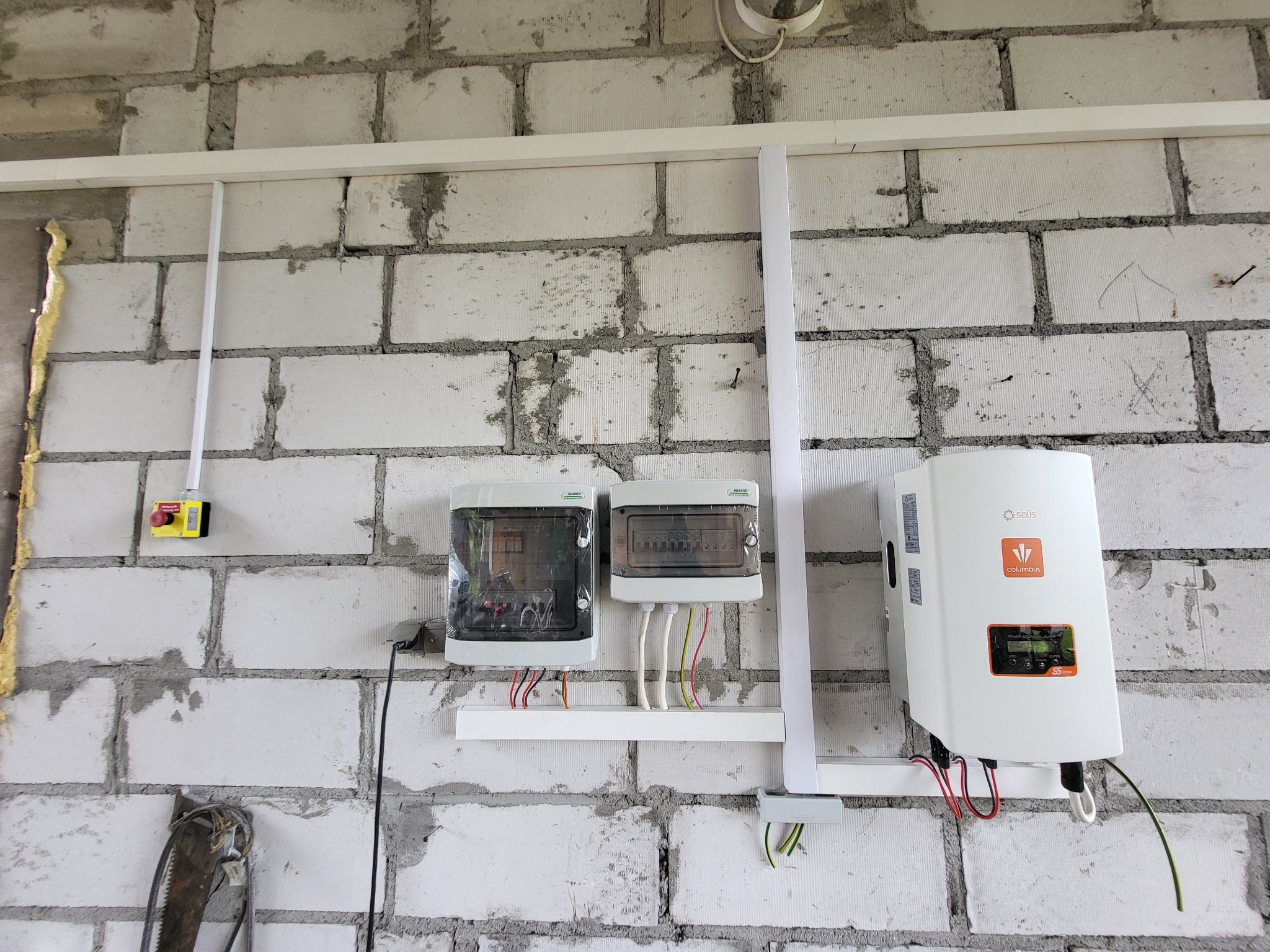 Instalacja elektryczna w surowym wnętrzu: dwa rozdzielnice, wyłącznik awaryjny i inwerter fotowoltaiczny Solis Columbus na białej ścianie z cegły.