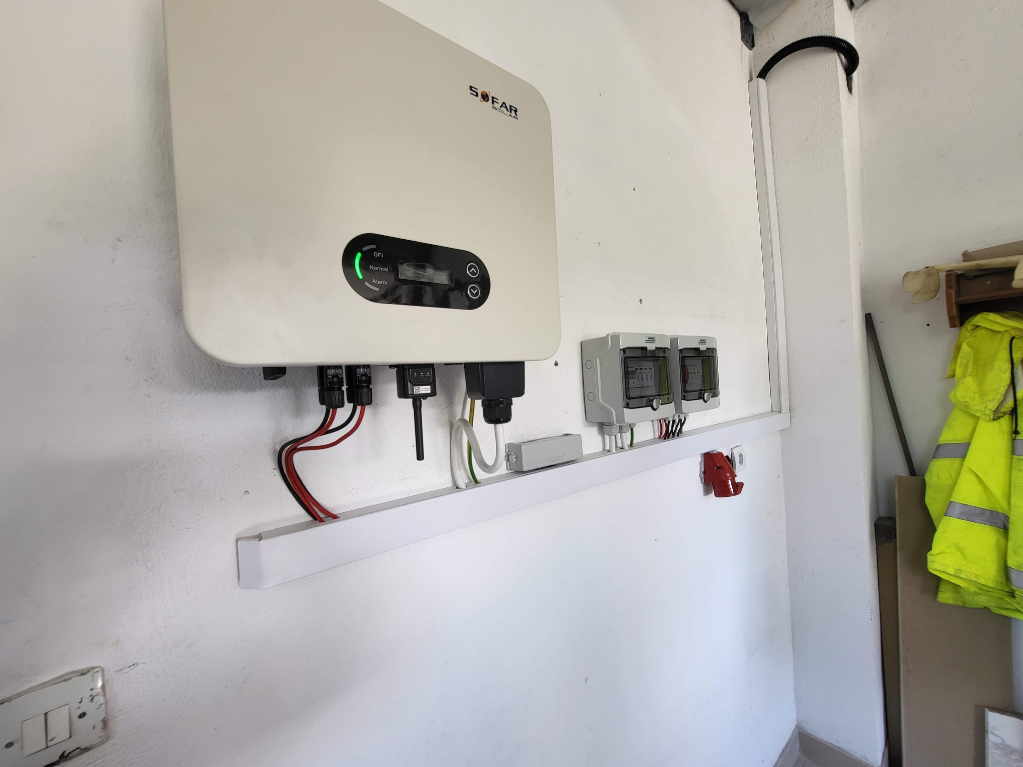 Biały inwerter SOFAR z zieloną diodą LED, zamontowany na ścianie z przewodami, obok dwa liczniki energii i gniazdo siłowe. Widoczna kurtka robocza.