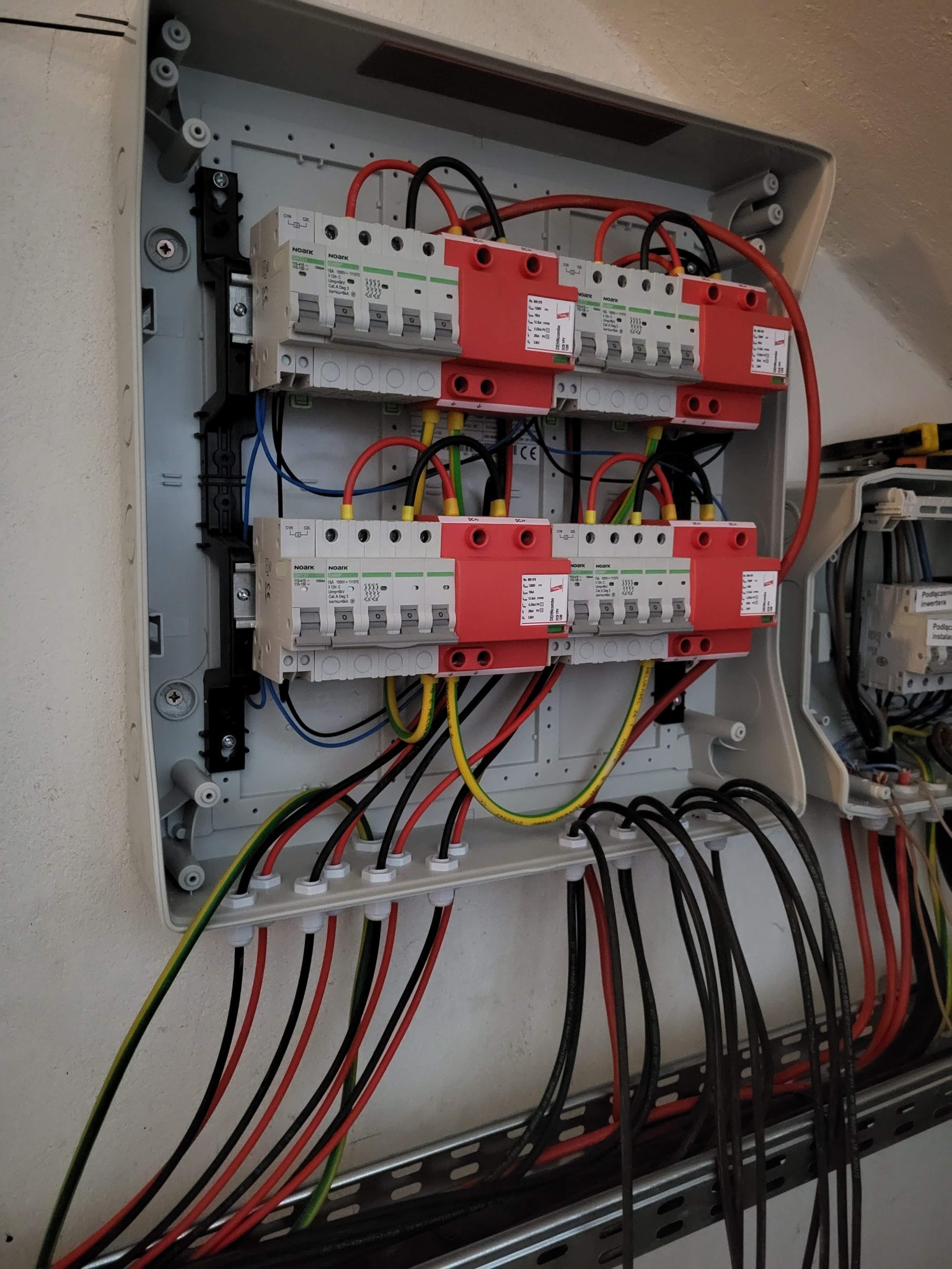 Rozdzielnica elektryczna z zabezpieczeniami przepięciowymi Noark, widoczne kable w kolorach: czarnym, czerwonym, żółto-zielonym. Instalacja w szarej obudowie.