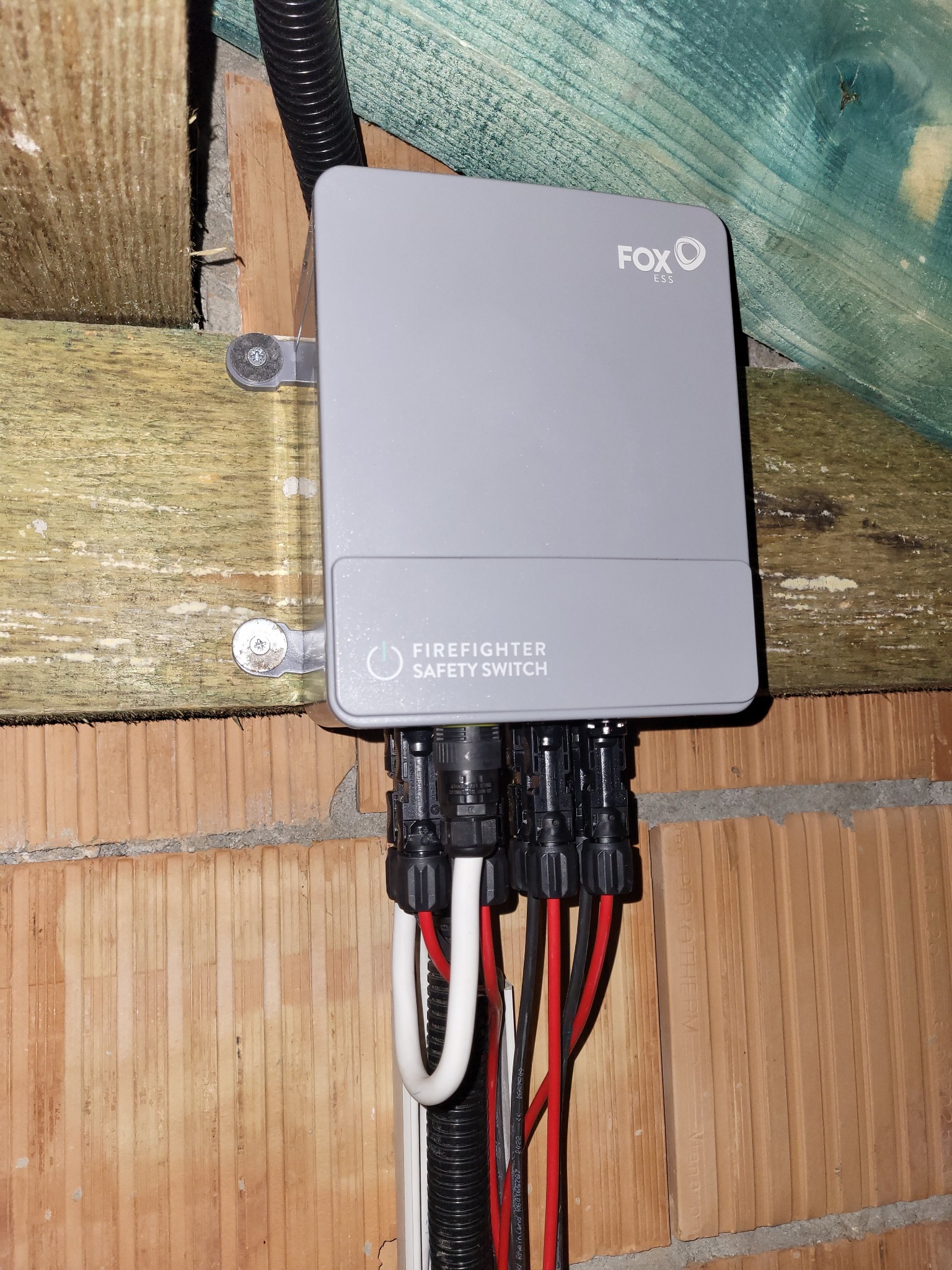 Szary wyłącznik bezpieczeństwa pożarowego marki Fox ESS, zamontowany na drewnianej konstrukcji i ceglanej ścianie, z widocznymi kablami i złączkami.