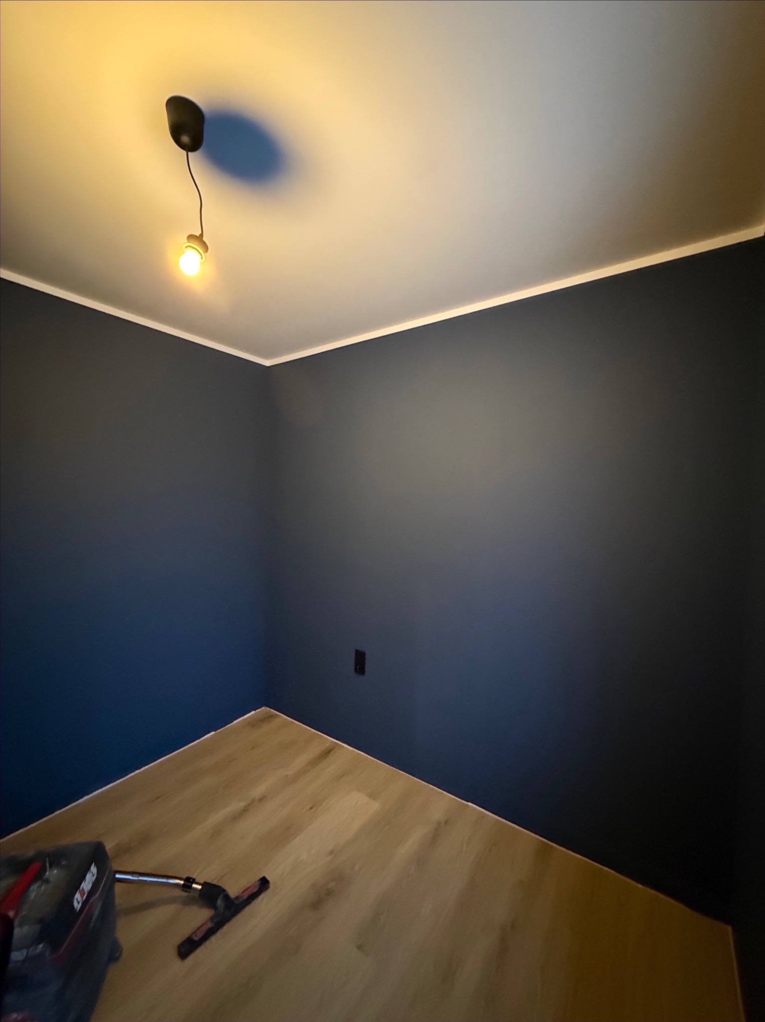 Pomieszczenie po malowaniu ścian na ciemny granat, z widoczną lampą sufitową i fragmentem podłogi z paneli. W rogu narzędzia malarskie.