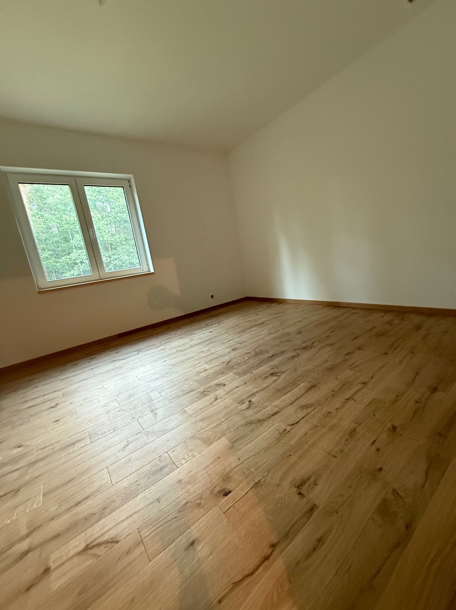 Jasne panele podłogowe w nowym pokoju z oknem, widok na zieleń za oknem. Świeżo pomalowane ściany w odcieniach bieli. Minimalistyczny styl wykończenia wnętrza.