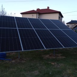 JM Instalacje Justyna Granat - Panel fotowoltaiczny na metalowej konstrukcji, z niebieskim przewodem w tle, na trawiastym terenie przed budynkiem z czerwoną dachówką. Częściowo widoczny samochód dostawczy.