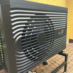 JM Instalacje Justyna Granat - Czarna pompa ciepła Fox Air na metalowej podstawie, ustawiona na kostce brukowej, z żółtą ścianą w tle. Widoczny wentylator i detale konstrukcyjne.