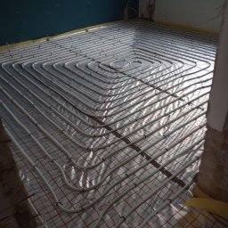 JM Instalacje Justyna Granat - Białe rury ogrzewania podłogowego ułożone spiralnie na izolacji z czerwoną siatką w surowym wnętrzu. Widoczne fragmenty ściany w kolorze ciemnego turkusu i okno.