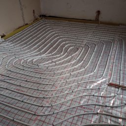 JM Instalacje Justyna Granat - Białe rury ogrzewania podłogowego ułożone spiralnie na folii z czerwoną siatką, gotowe do zalania posadzką. Widoczne fragmenty ścian i przewodów elektrycznych.