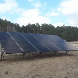 Panel fotowoltaiczny na tle lasu i nieba z chmurami, zamontowany na metalowej konstrukcji. Obok leży karton, widoczny kabel. Ujęcie z ziemi.