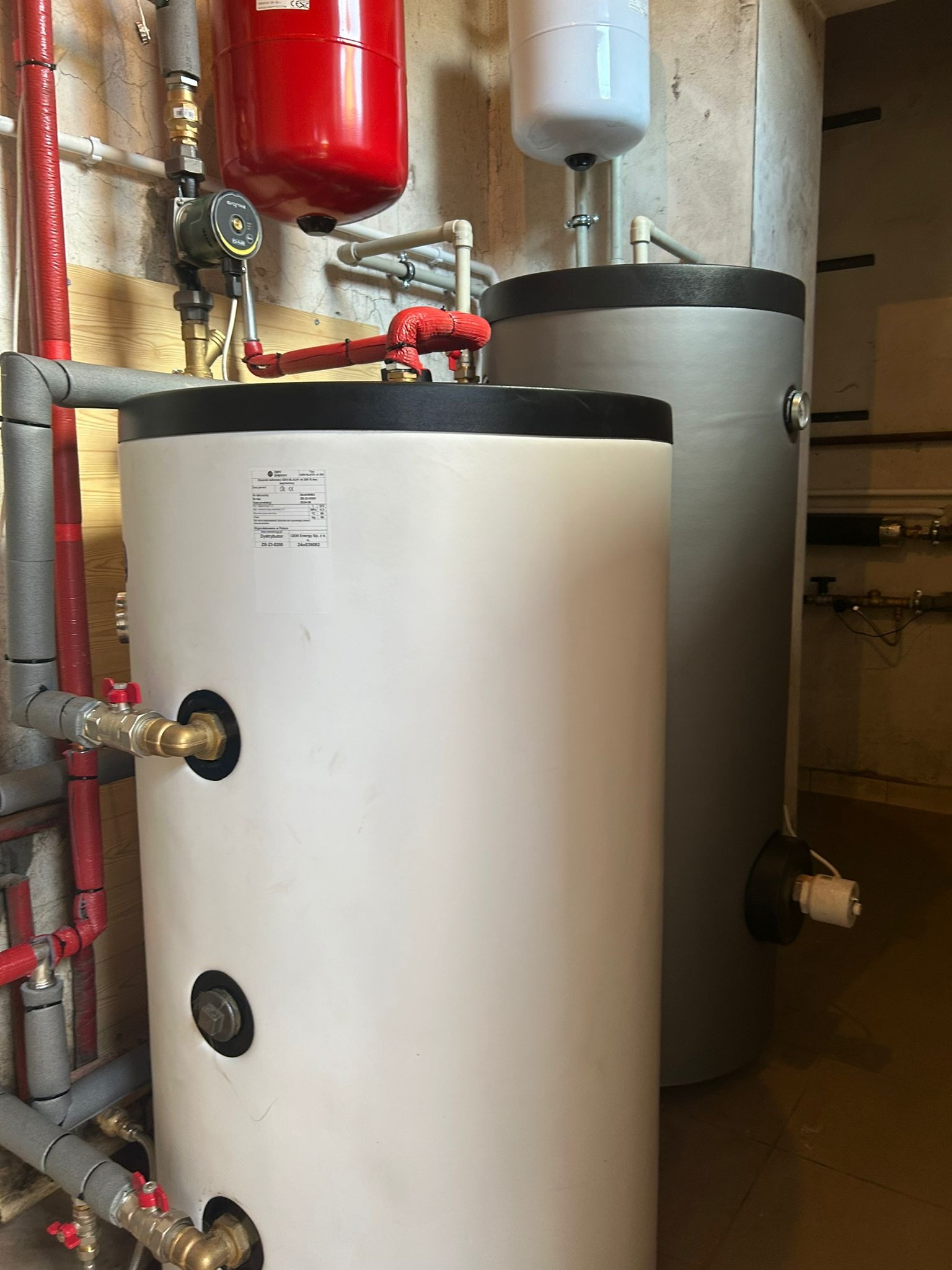 Dwa zbiorniki akumulacyjne, biały i szary, z instalacją hydrauliczną w kotłowni. Widoczne rury, zawory i manometry. Elementy instalacji grzewczej, oświetlenie górne.