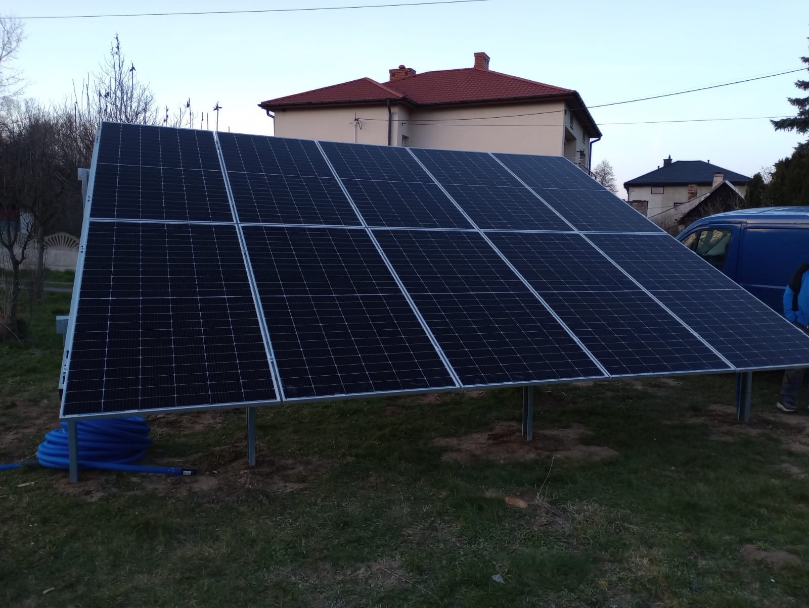 Panel fotowoltaiczny na metalowej konstrukcji, z niebieskim przewodem w tle, na trawiastym terenie przed budynkiem z czerwoną dachówką. Częściowo widoczny samochód dostawczy.