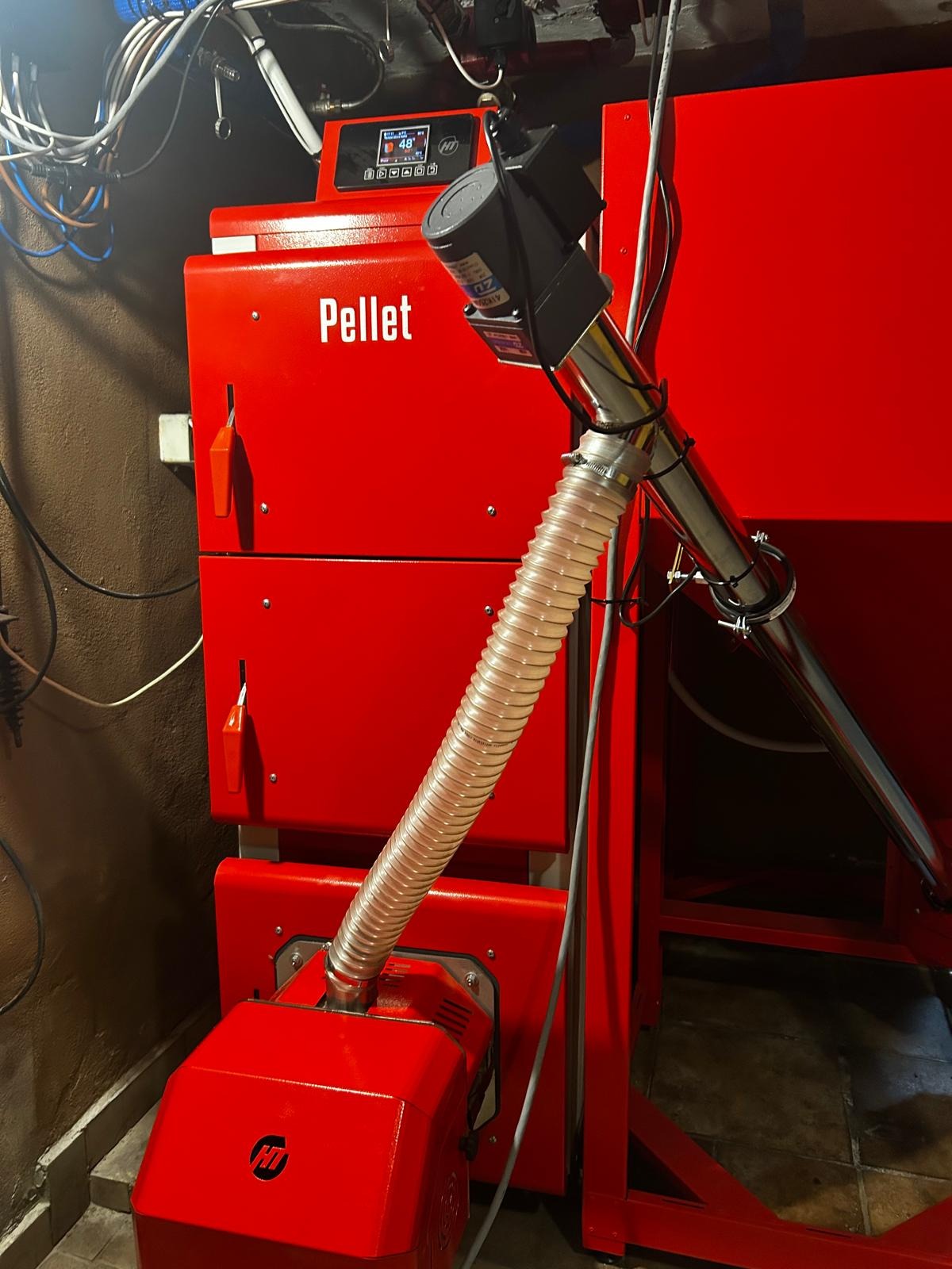 Czerwony piec na pellet z podajnikiem, widoczne przewody i rury. Na górze panel sterowania z wyświetlaczem. Oświetlenie sufitowe i fragment podłogi w tle.