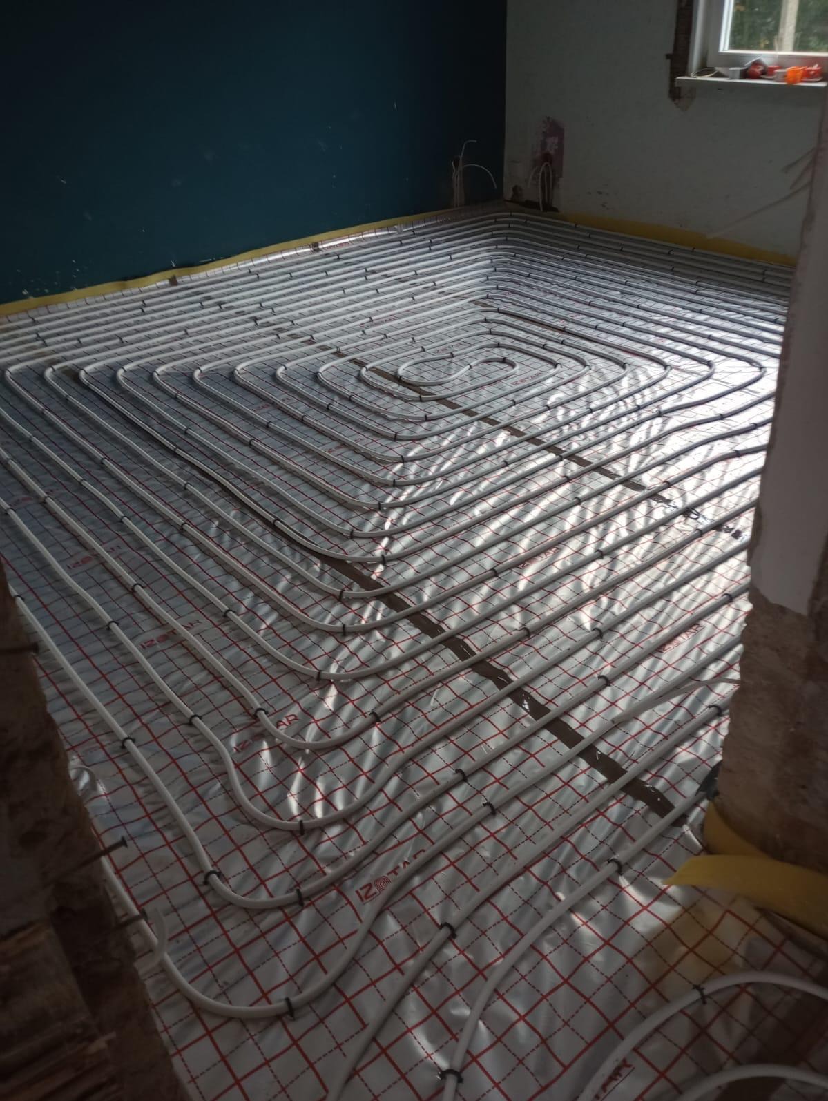 Białe rury ogrzewania podłogowego ułożone spiralnie na izolacji z czerwoną siatką w surowym wnętrzu. Widoczne fragmenty ściany w kolorze ciemnego turkusu i okno.