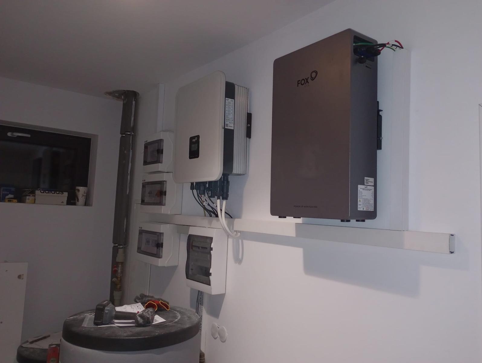 Zestaw paneli fotowoltaicznych FOX ESS zamontowany na ścianie wewnątrz pomieszczenia. Widoczne przewody i skrzynki elektryczne. Na pierwszym planie zbiornik.