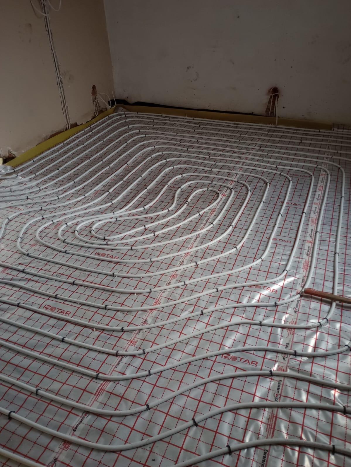 Białe rury ogrzewania podłogowego ułożone spiralnie na folii z czerwoną siatką, gotowe do zalania posadzką. Widoczne fragmenty ścian i przewodów elektrycznych.