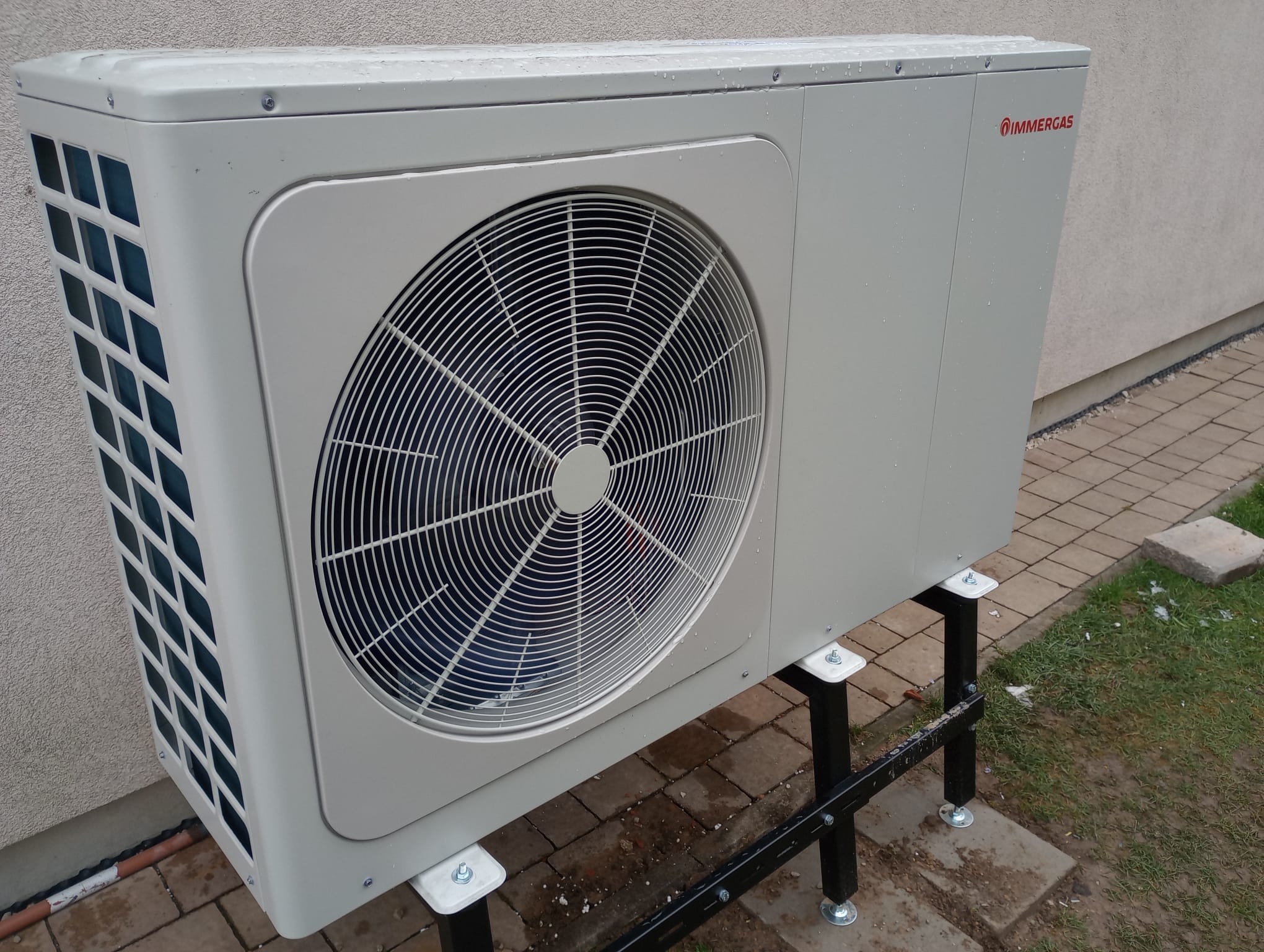 Biała pompa ciepła Immergas na czarnym stelażu, ustawiona na zewnątrz budynku. Widoczne detale konstrukcyjne i wentylator. Zdjęcie z poziomu gruntu, lekko z dołu.
