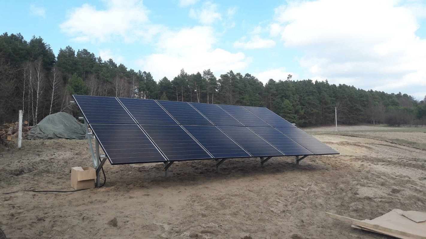 Panel fotowoltaiczny na tle lasu i nieba z chmurami, zamontowany na metalowej konstrukcji. Obok leży karton, widoczny kabel. Ujęcie z ziemi.
