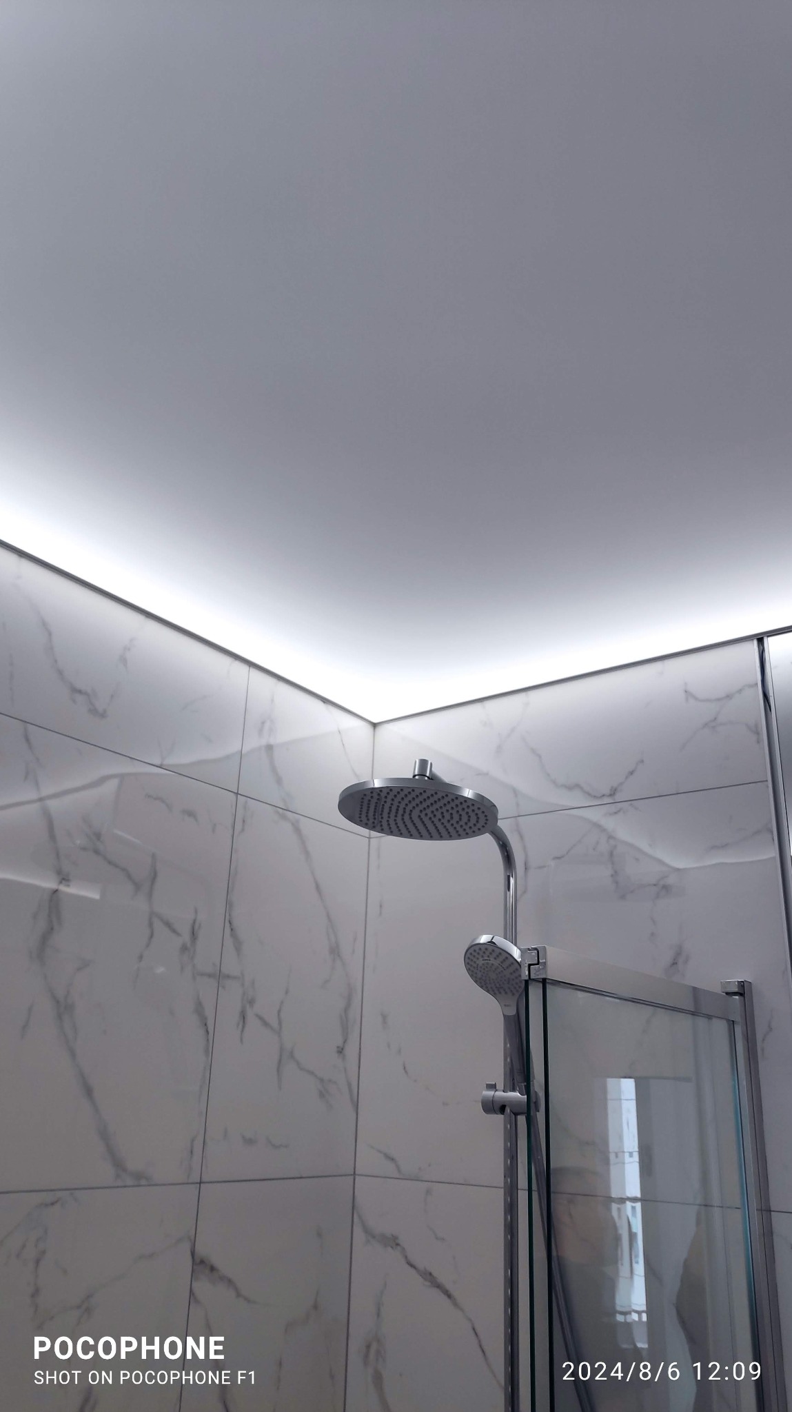 Nowoczesna kabina prysznicowa z deszczownicą i marmurowymi płytkami, oświetlona listwą LED pod sufitem. Minimalistyczny design łazienki.