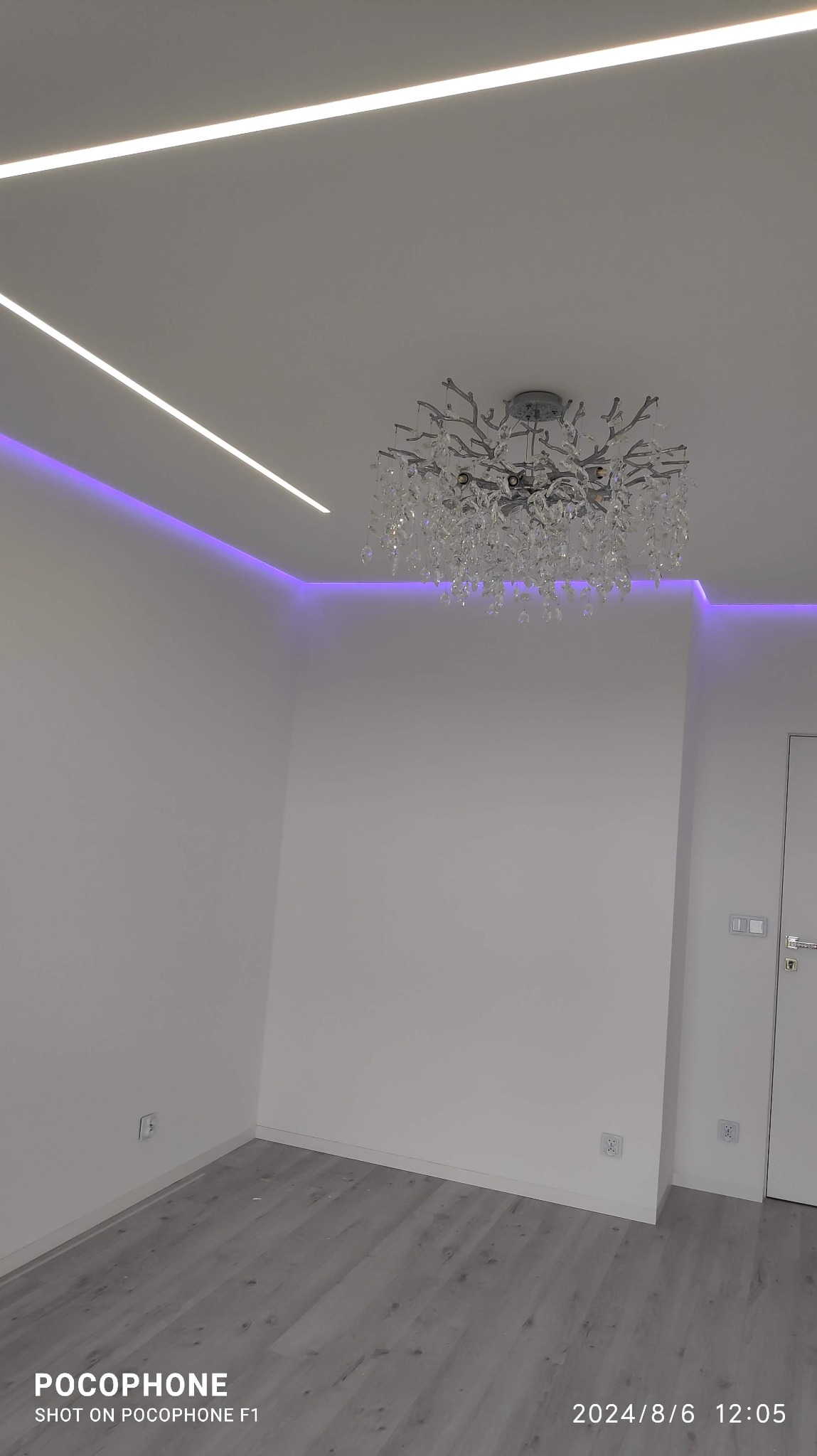 Białe wnętrze z nowoczesnym oświetleniem LED i kryształowym żyrandolem. Widoczne listwy LED w suficie i subtelne fioletowe podświetlenie. Szare panele podłogowe.