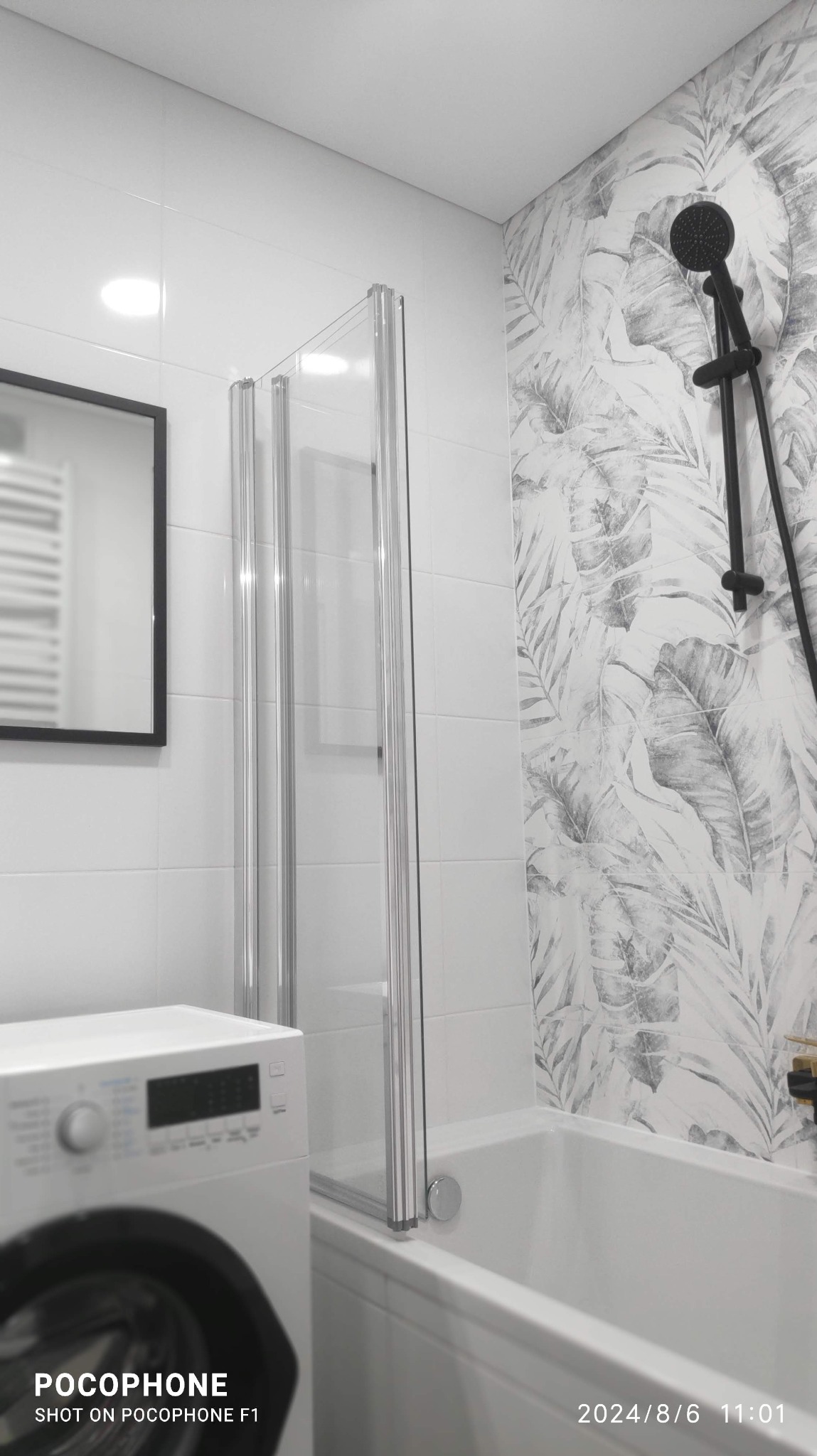 Nowoczesna łazienka z wanną i czarną armaturą, kabina prysznicowa ze srebrnymi profilami, wzorzyste płytki na ścianie. Widoczna pralka i lustro.