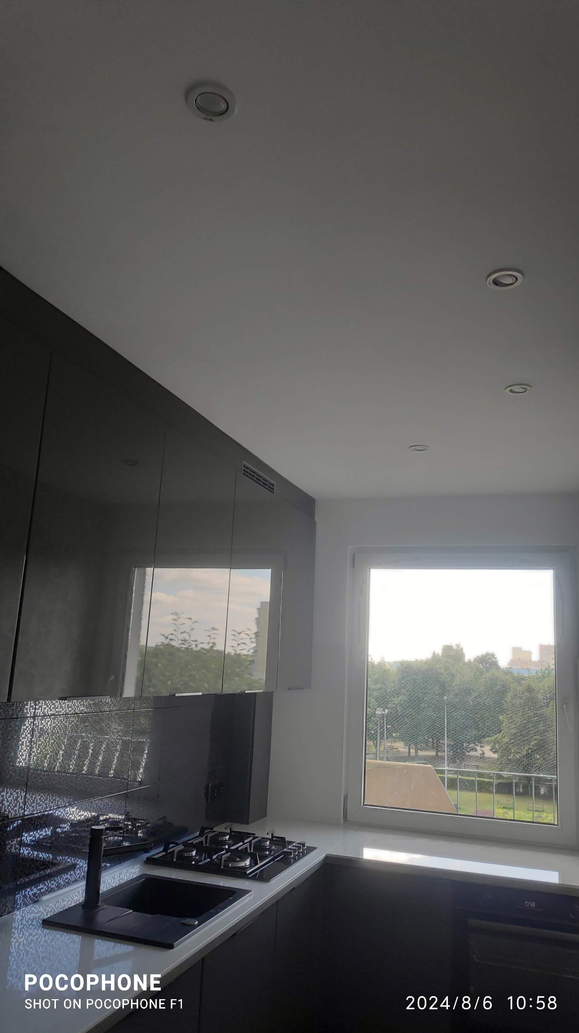 Nowoczesna kuchnia z czarnymi, błyszczącymi szafkami, blatem i płytą grzewczą. Widok na zieleń za oknem. Oświetlenie sufitowe punktowe.