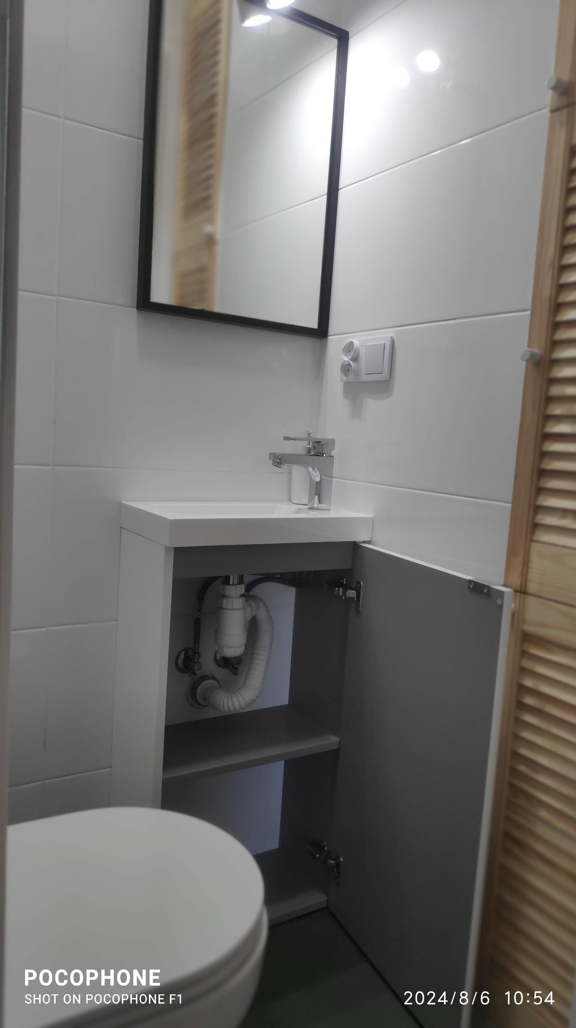 Biała łazienka z umywalką w szafce z otwartymi drzwiczkami, widoczne rury odpływowe. Nad umywalką lustro w czarnej ramie i gniazdko elektryczne.