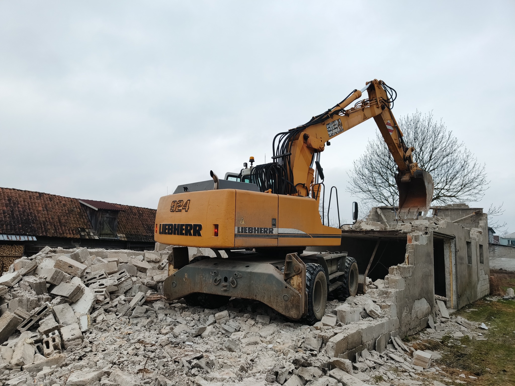 Żółta koparka marki Liebherr na gruzowisku, w tle budynek w trakcie rozbiórki, pochmurne niebo. Widoczny fragment dachu budynku gospodarczego.