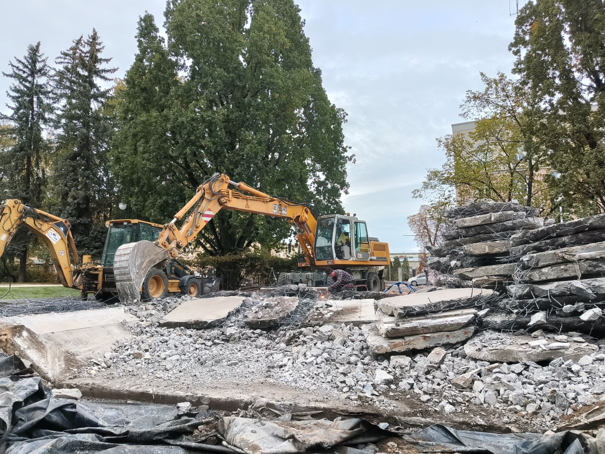 Żółte koparki i gruzowisko po rozbiórce, z widocznym spawaczem przy pracy. W tle zieleń drzew i budynek. Ujęcie z bliska, ukazujące detale gruzu i sprzętu.