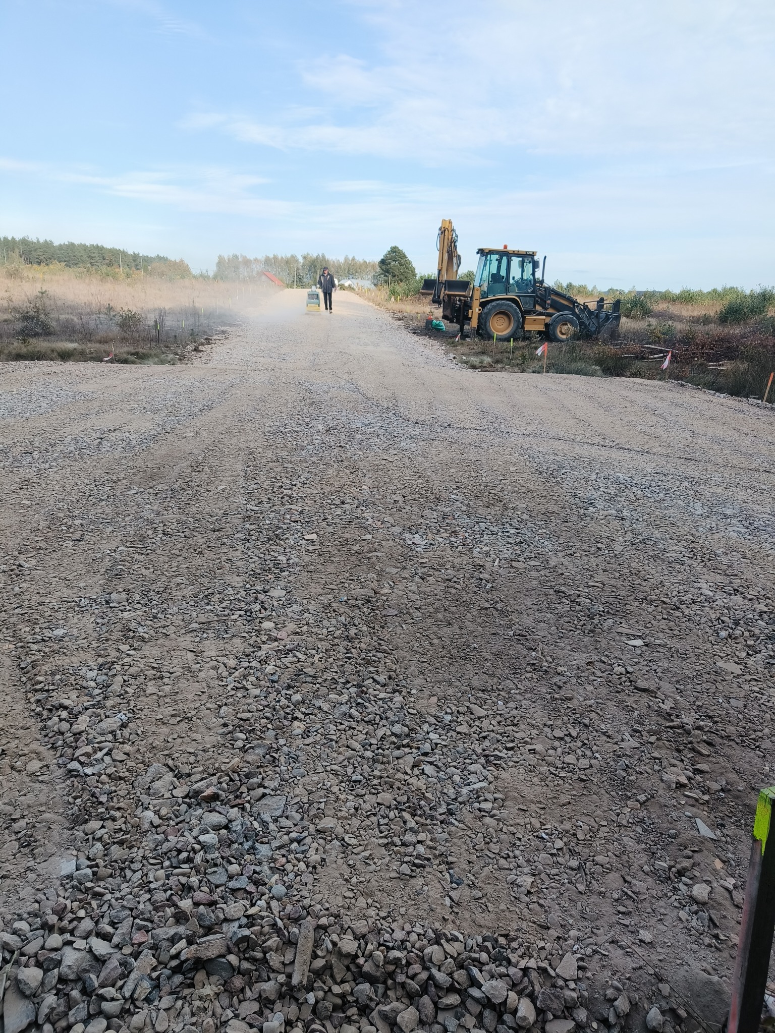 Utwardzona droga gruntowa z rozsypanym kamieniem, równana przez pracownika z maszyną. W tle las i błękitne niebo. Widoczny sprzęt budowlany.