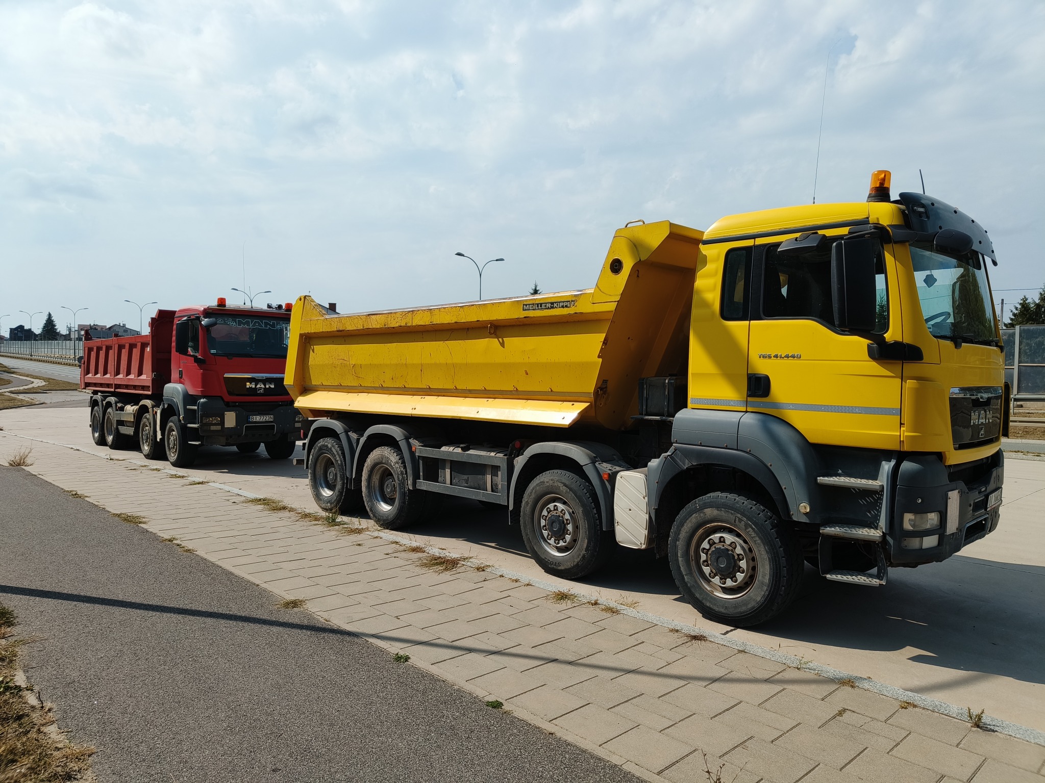 Żółta i czerwona wywrotka MAN w Białymstoku, zaparkowane obok siebie na chodniku. Pojazdy gotowe do transportu materiałów budowlanych.