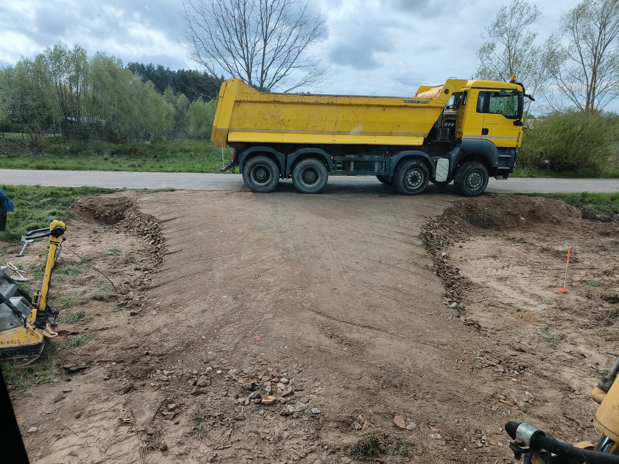 Żółty wywrotka na drodze w trakcie utwardzania, widoczna ubijarka i wykopany fragment drogi. Prace ziemne w toku, krajobraz wiejski w tle.