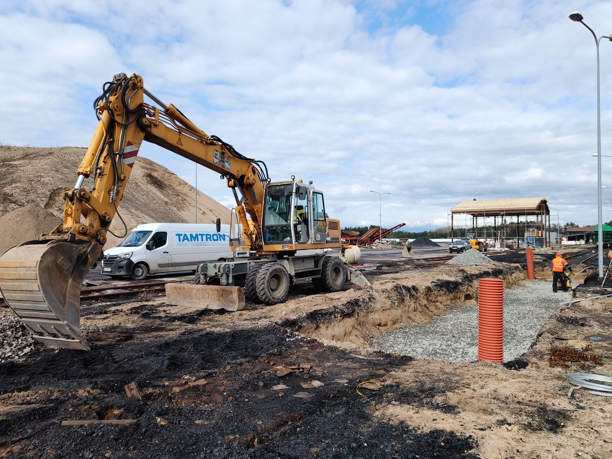 Żółta koparka kołowa Liebherr 924, wykop z podsypką żwirową i rurą drenarską w ziemi, biały bus Tamtron w tle, błękitne niebo z chmurami.