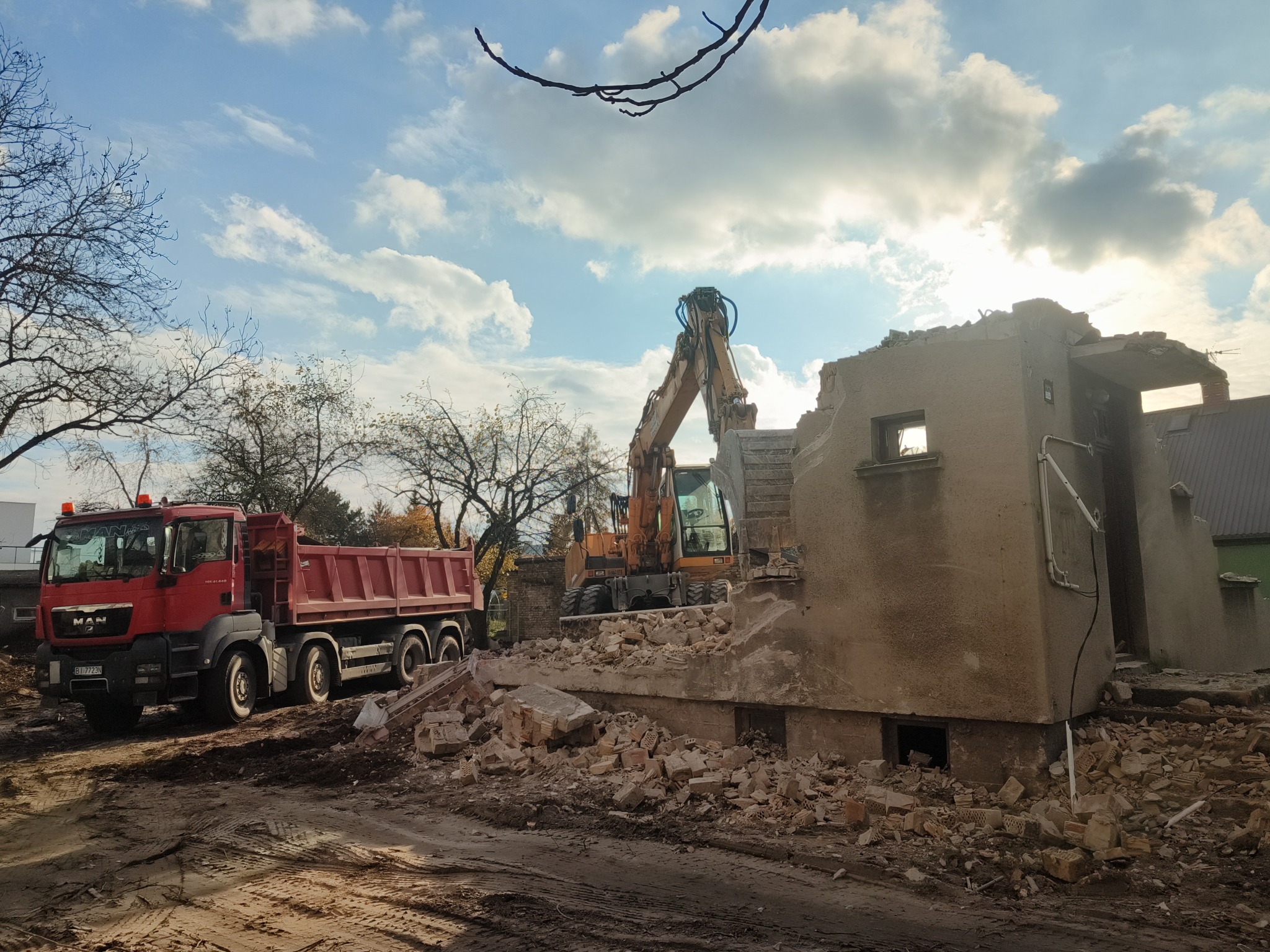 Ciężki sprzęt (koparka i wywrotka MAN) przy pozostałościach budynku w trakcie rozbiórki. Gruz i fragmenty ścian w otoczeniu. Błękitne niebo w tle.