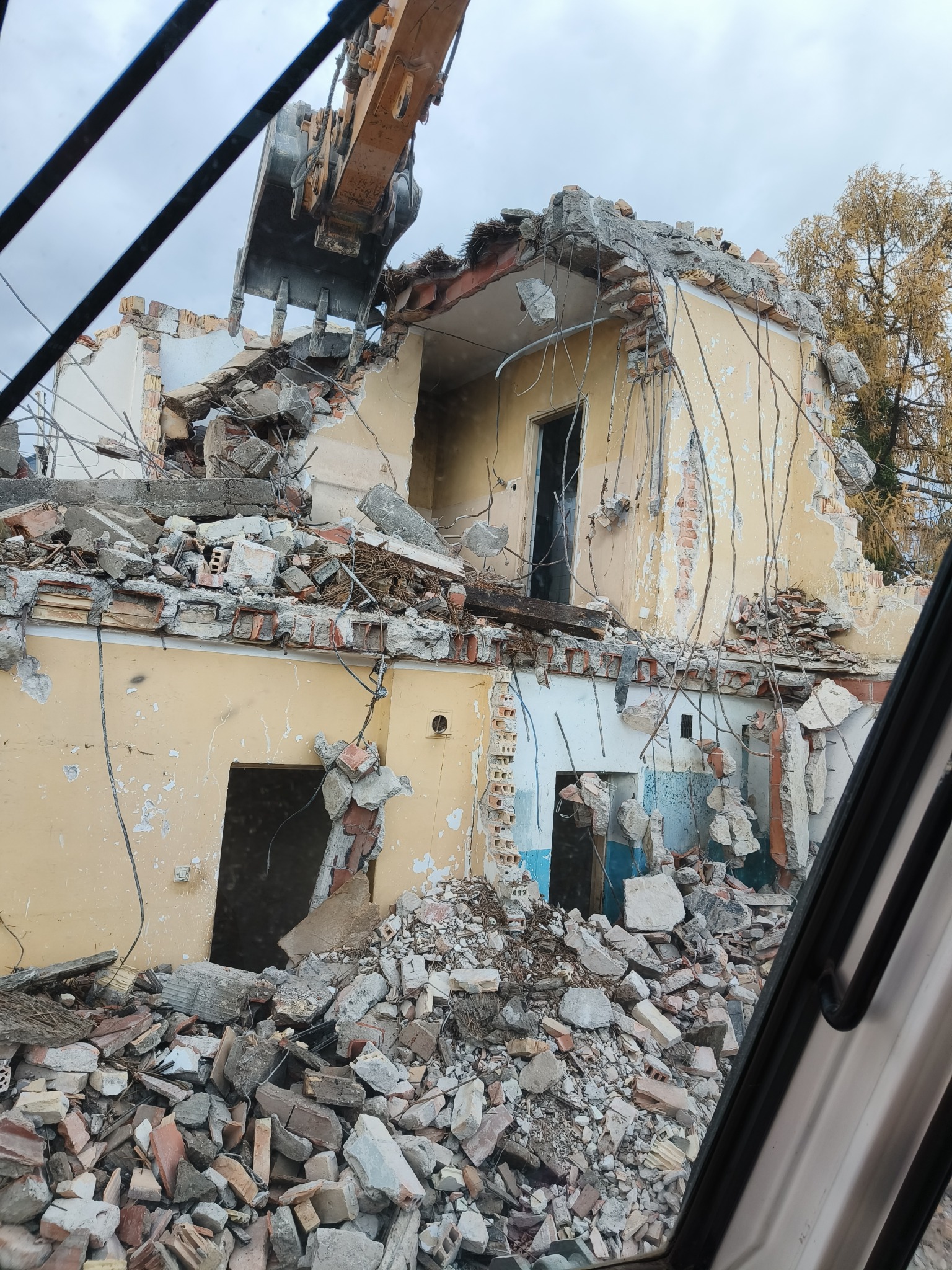 Żółty budynek w trakcie rozbiórki przez koparkę z łyżką z zębami, widok z wnętrza kabiny. Gruz i odsłonięte cegły.