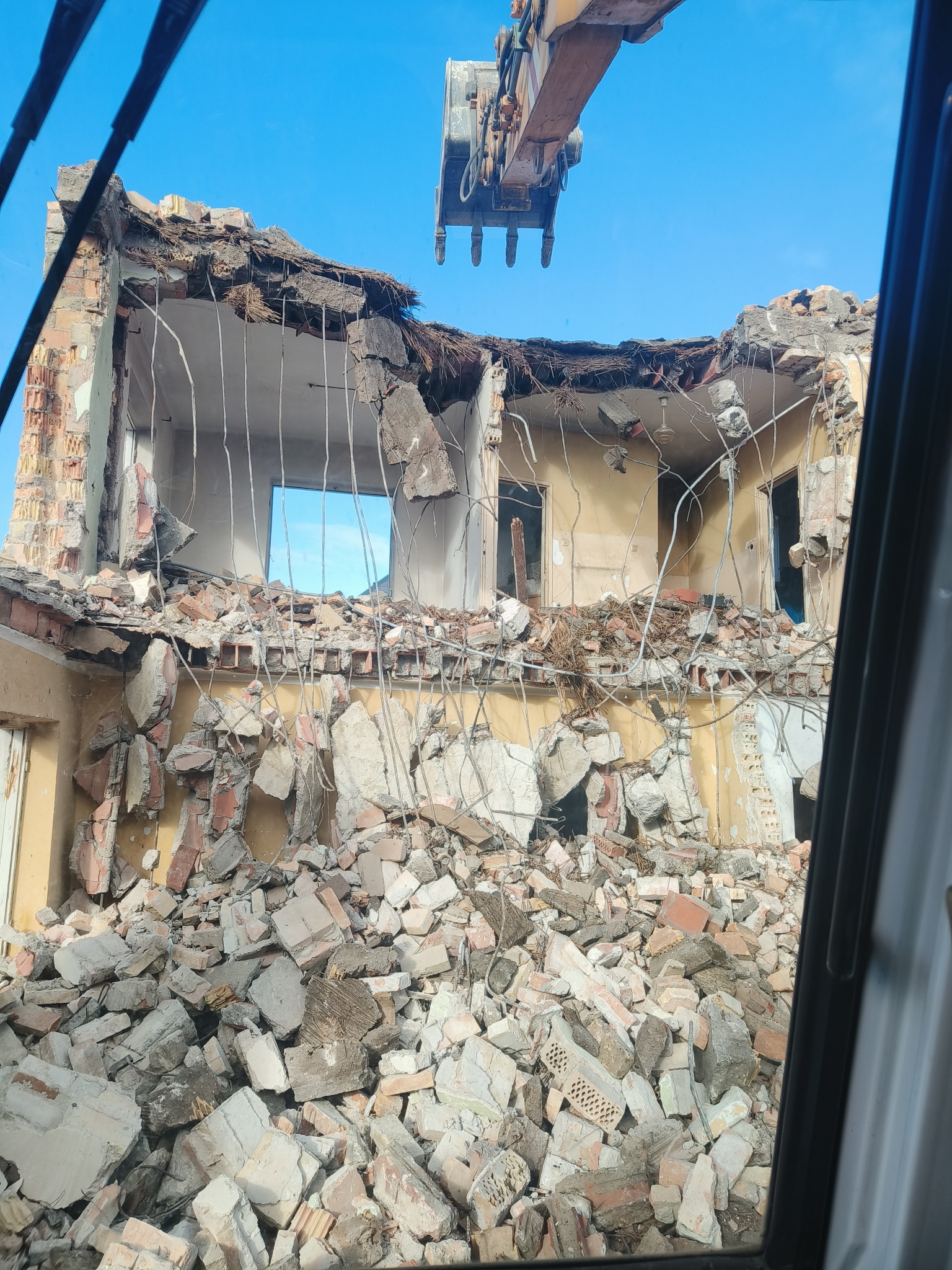 Ekskawator w trakcie rozbiórki budynku z cegły, widok z wnętrza kabiny. Gruz i fragmenty ścian w dole kadru, błękitne niebo w tle.