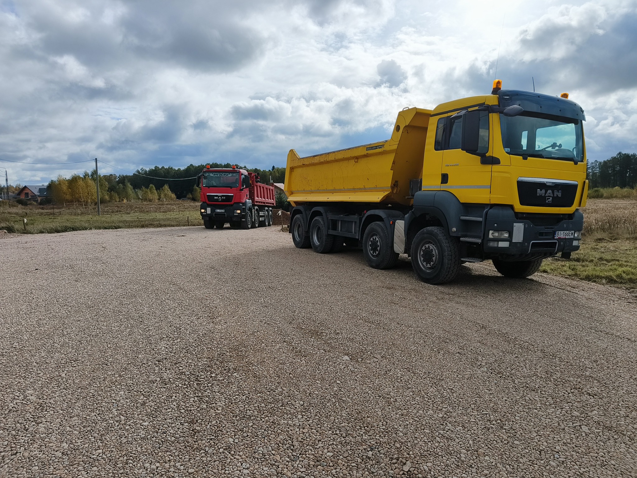 Żółta ciężarówka MAN na żwirowej drodze, w tle czerwona ciężarówka i pochmurne niebo. Widoczny etap utwardzania drogi.