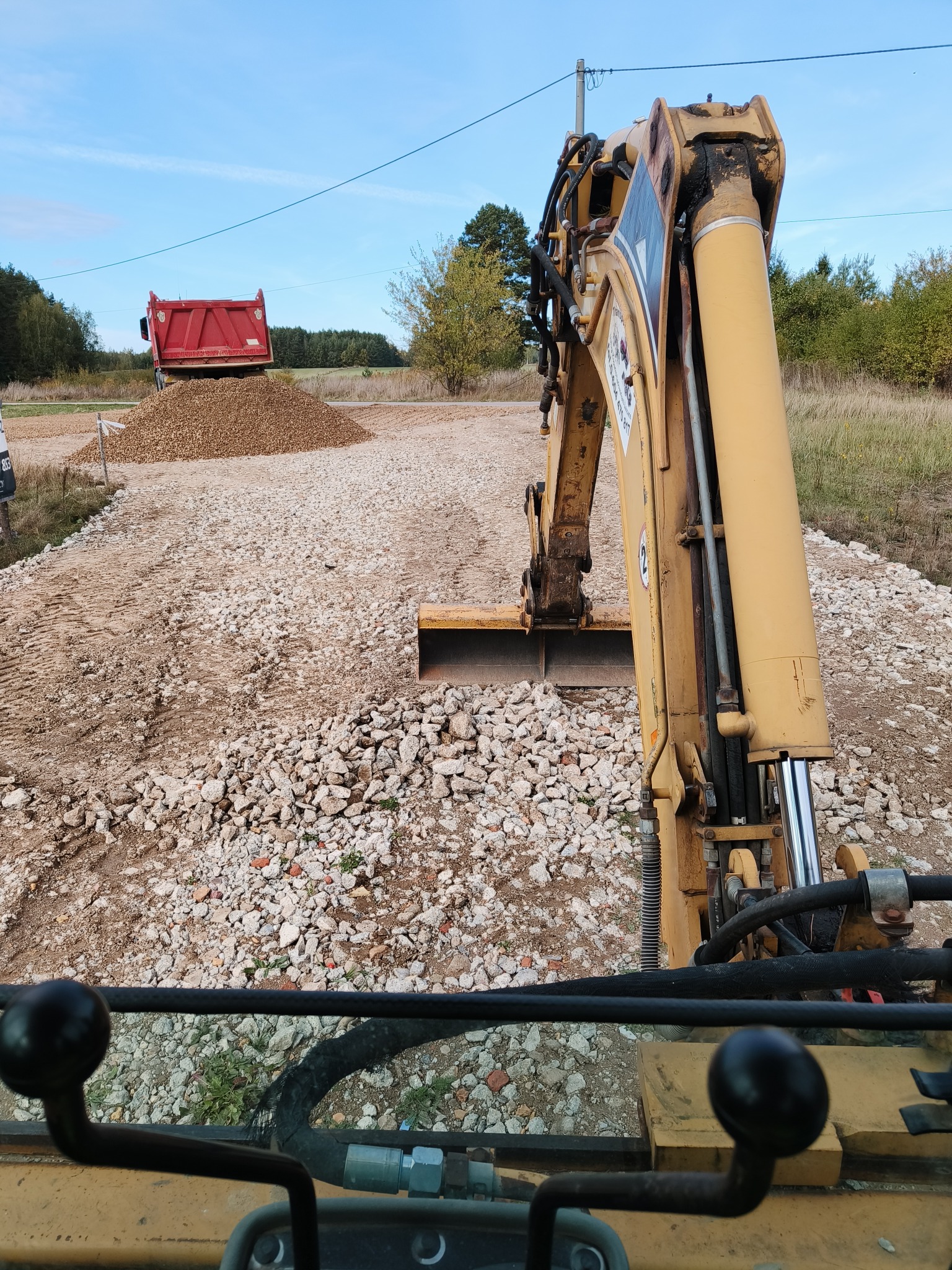 Żółta koparka na tle drogi z gruzem i czerwonej ciężarówki z górą żwiru. Widok z kabiny operatora. Utwardzanie drogi w toku.