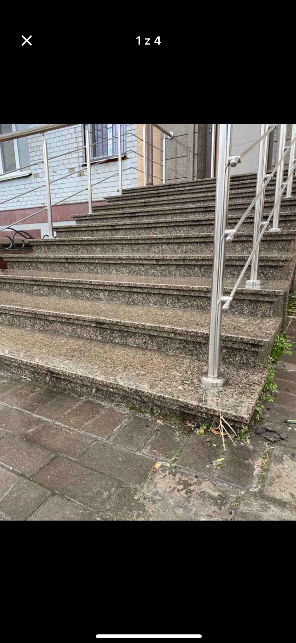 Zewnętrzne schody z jasnego kamienia z metalową balustradą. Widoczne płytki chodnikowe i fragment elewacji budynku. Ujęcie z dołu, podkreślające wysokość schodów.