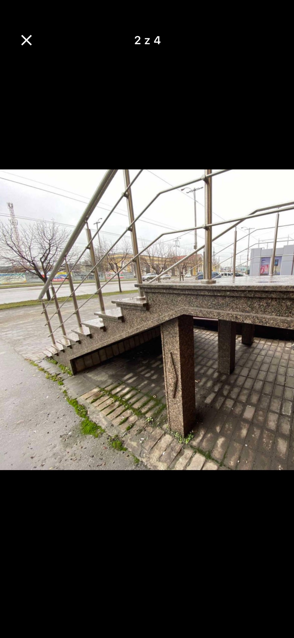 Zewnętrzne schody z szarego kamienia z metalową balustradą, prowadzące do budynku. Widoczne elementy konstrukcyjne i otoczenie miejskie w pochmurny dzień.