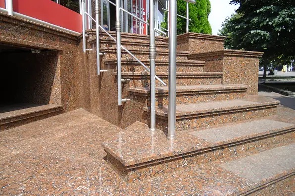 Zewnętrzne schody wejściowe wyłożone brązowym kamieniem z metalową balustradą. Widoczny fragment chodnika i wejście do piwnicy, również wykończone kamieniem.